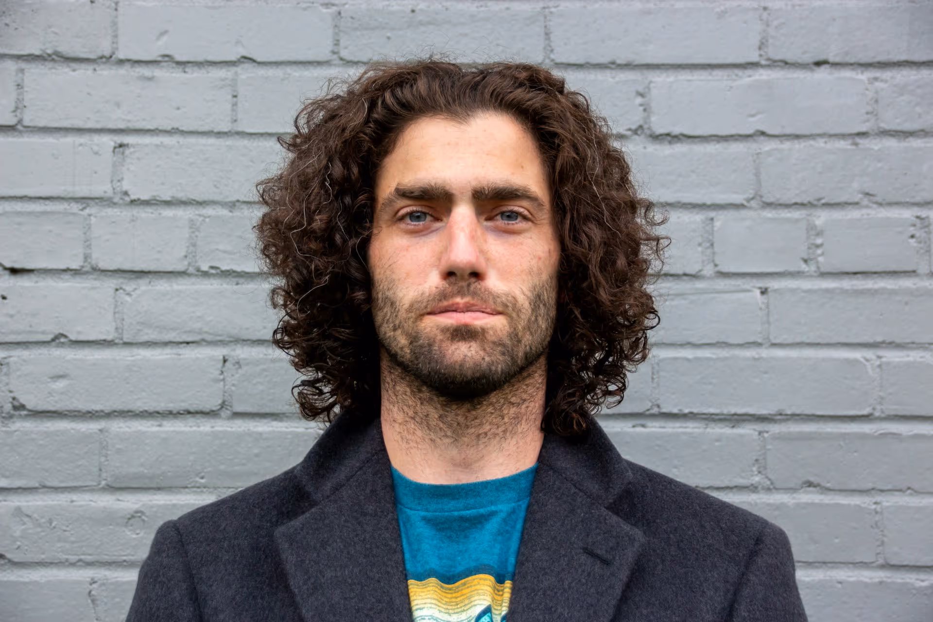 Man with curly hair, beard, and blue eyes wearing a coat over a blue shirt in front of a gray brick wall.