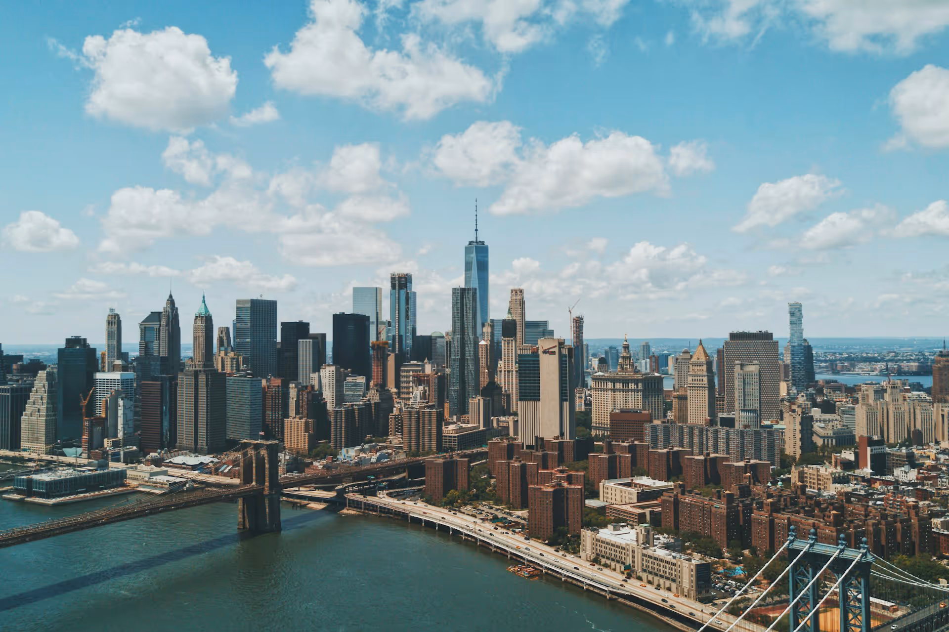 Aerial view of Manhattan skyline with Brooklyn Bridge over the East River on a partly cloudy day.