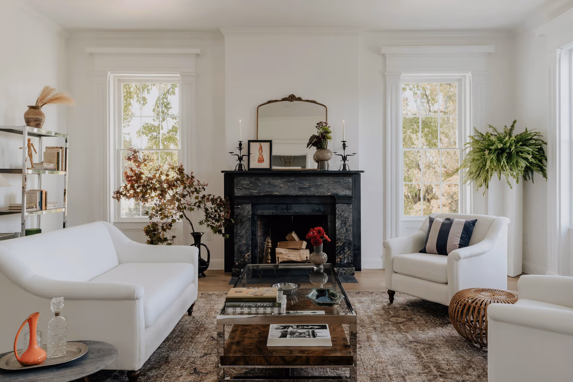 Bright living room with white sofas, glass coffee table, black fireplace, and large windows with greenery outside.