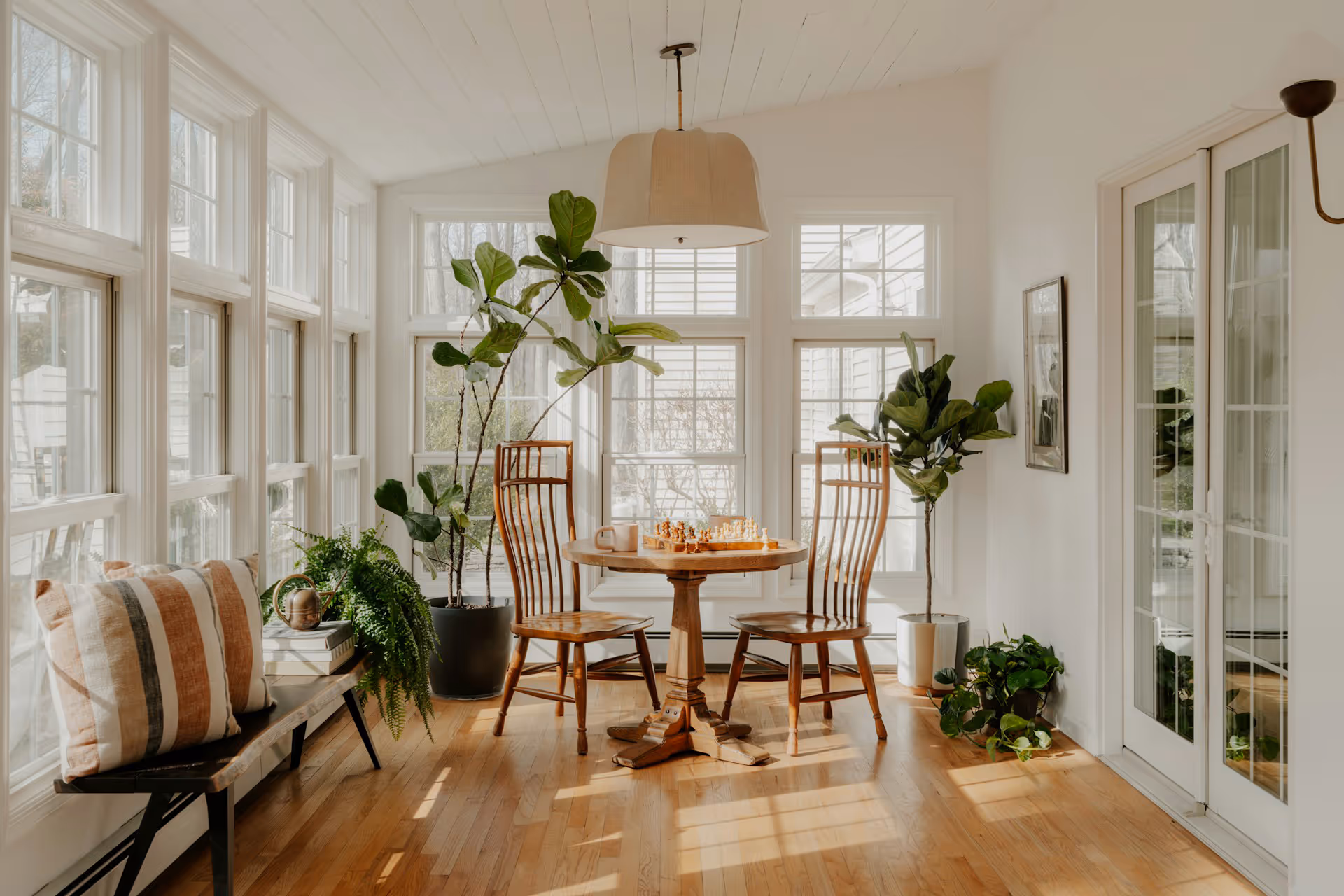 Sunlit room with wooden table and chairs, chessboard, plants, bench with striped pillows, and large windows.