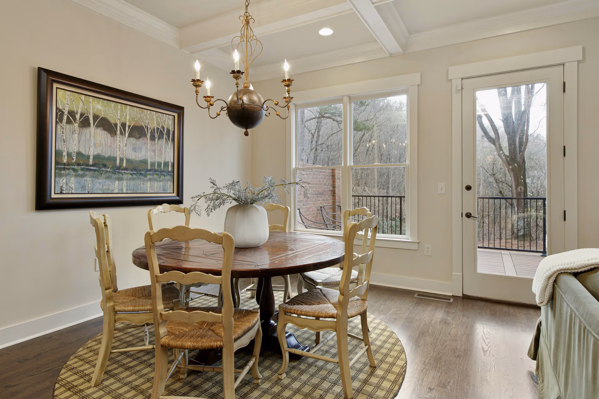 Dining area with round wooden table, six chairs, chandelier, and large window with outdoor view.