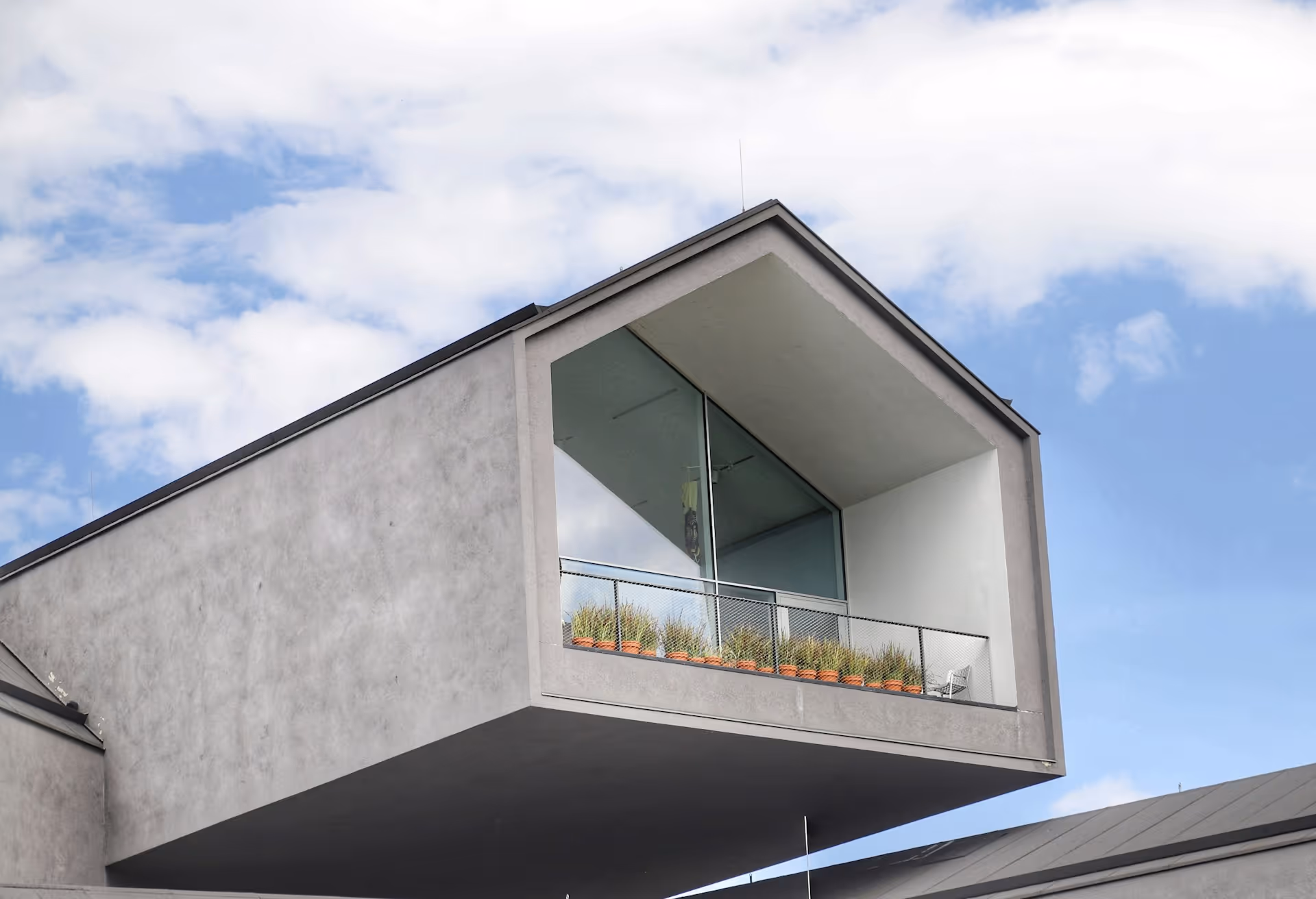 Modern concrete building with an elevated balcony featuring plants and a metal railing.