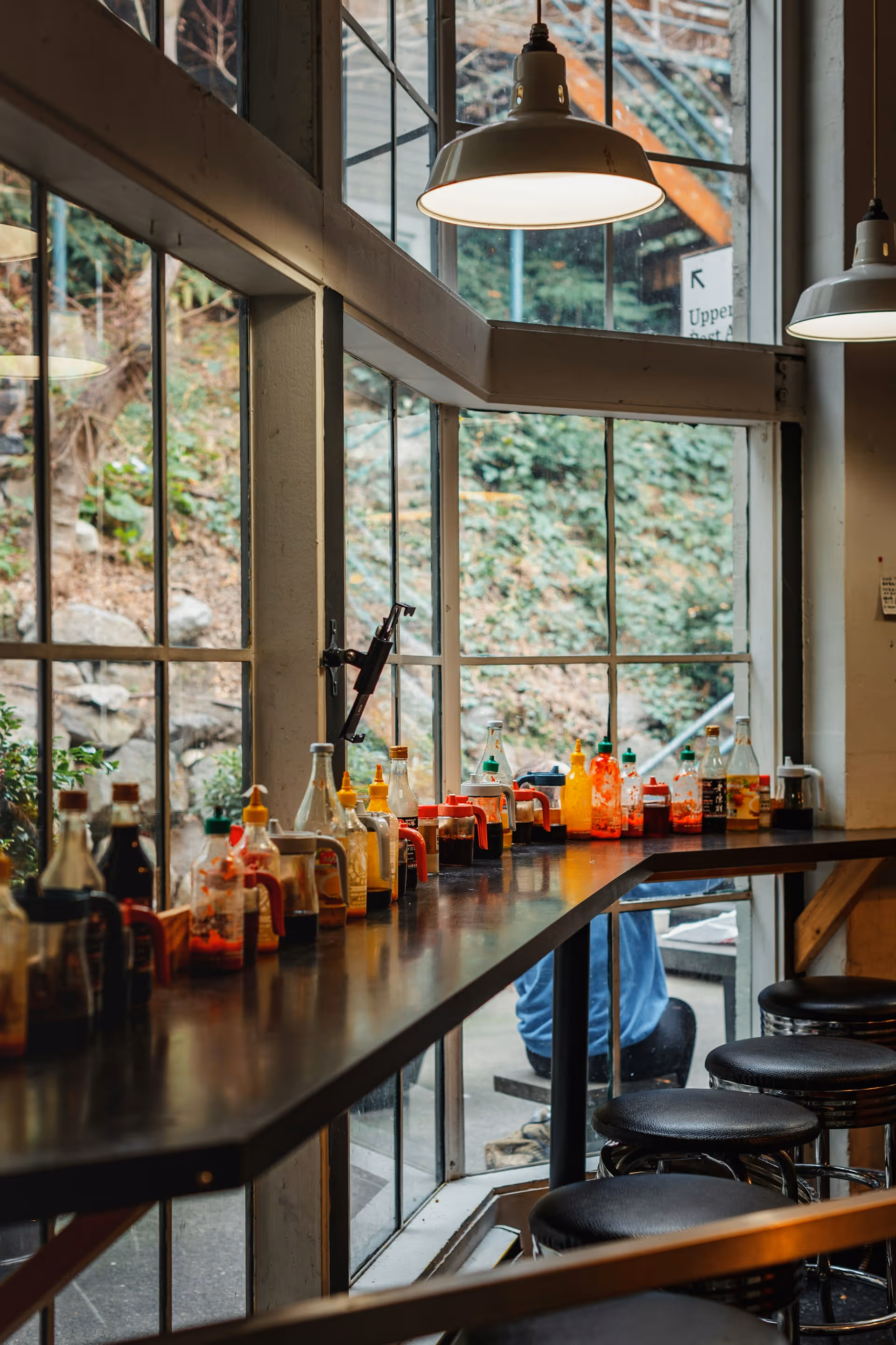 Counter lined with various condiment bottles and stools beside large windows in a cozy cafe.