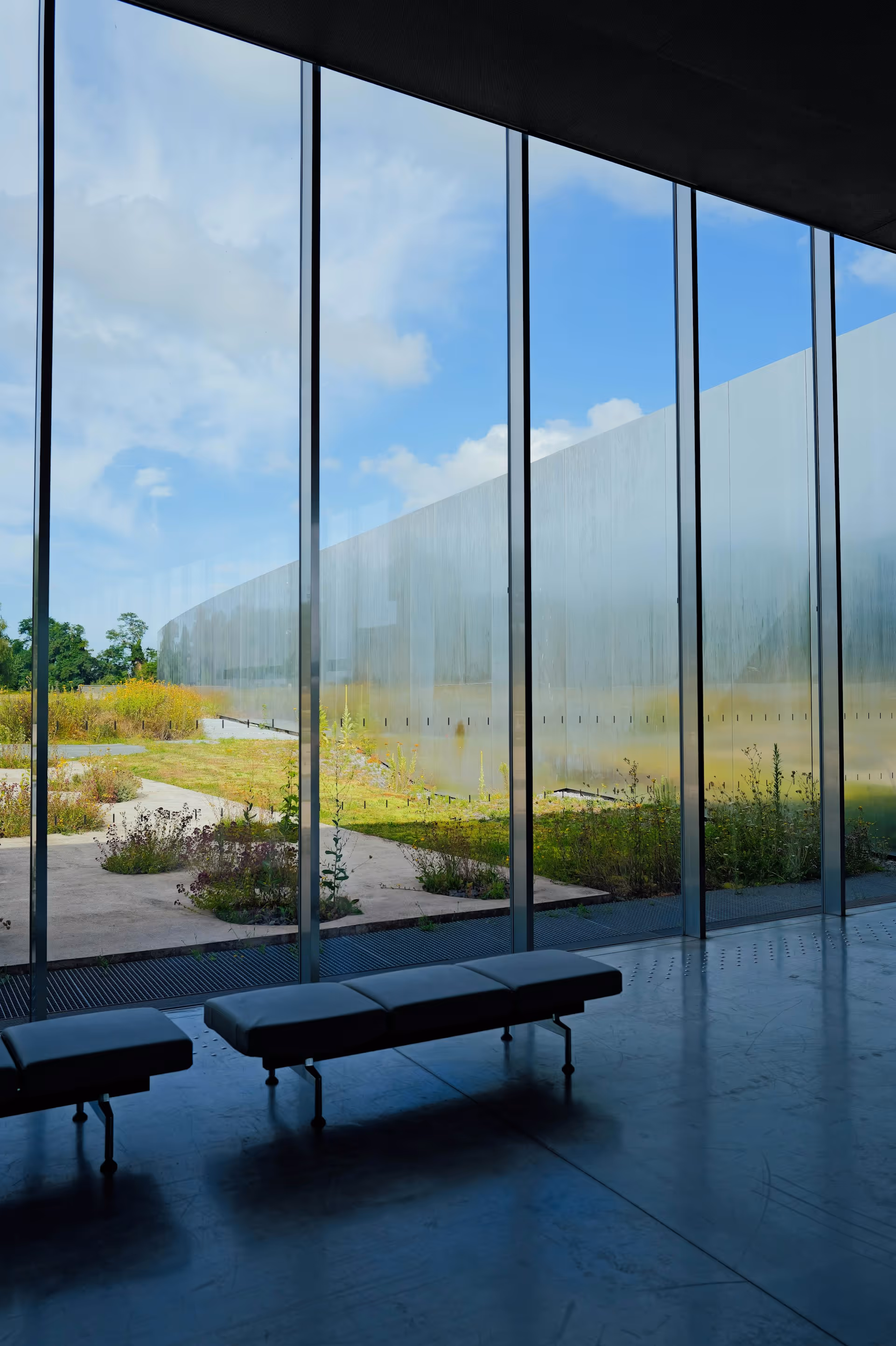 Modern interior with glass wall overlooking green garden and pathway outside under blue sky.