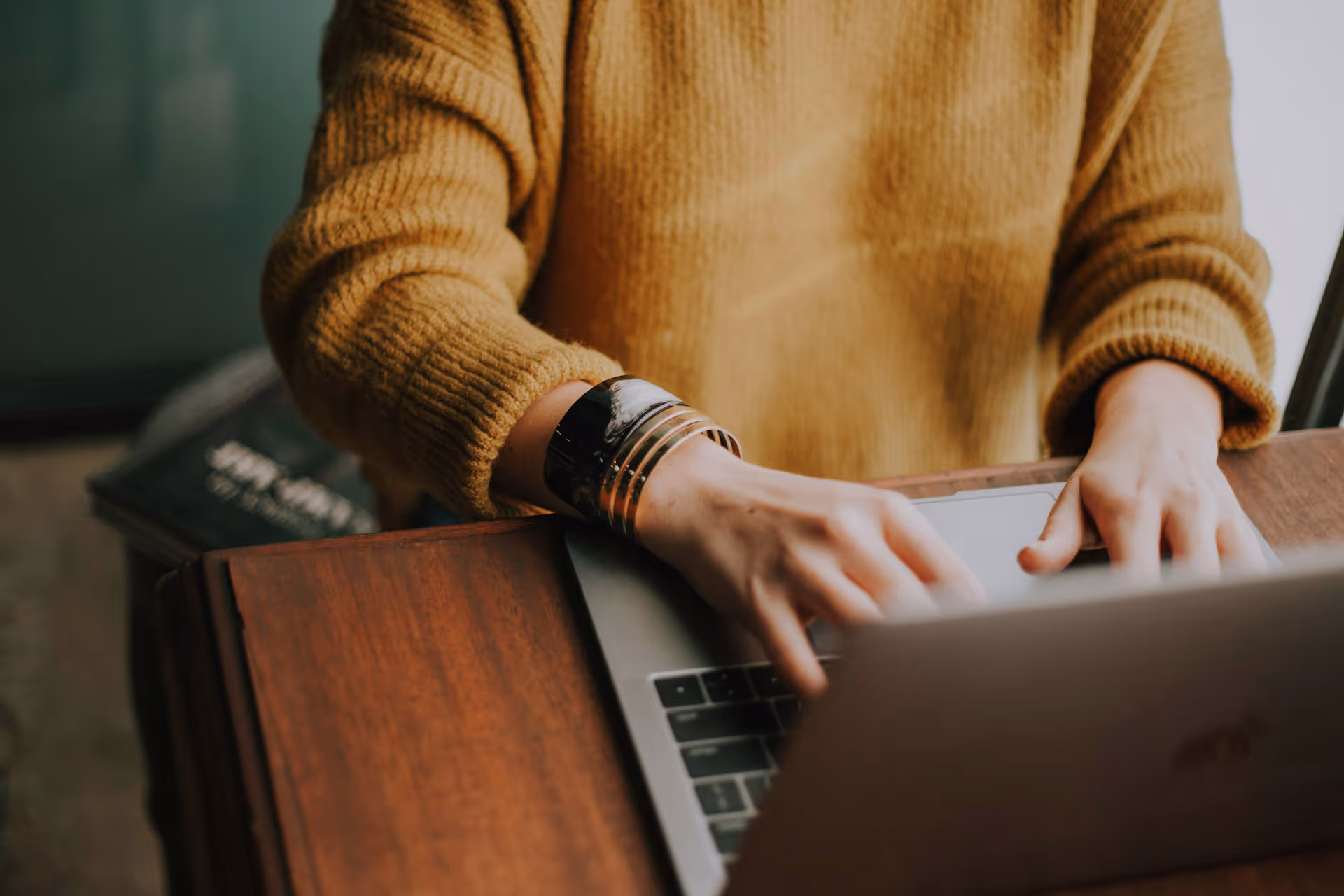 Person in brown sweater typing on a laptop at a wooden table with a book in background.