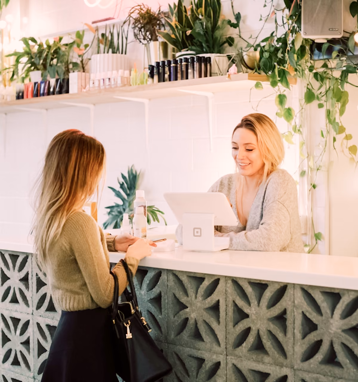 Customer at counter interacting with smiling staff in a bright shop with plants and beauty products.
