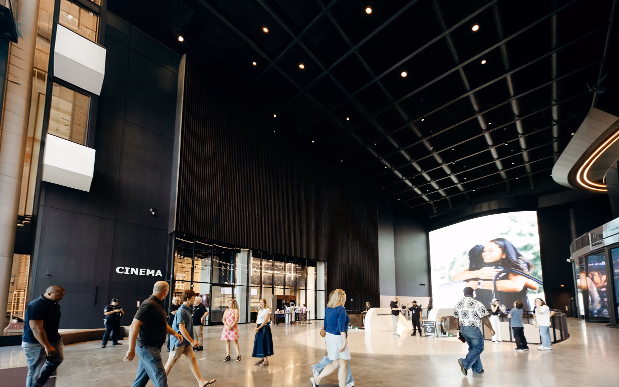 People walking and standing inside a modern cinema lobby with large digital screen and escalators.