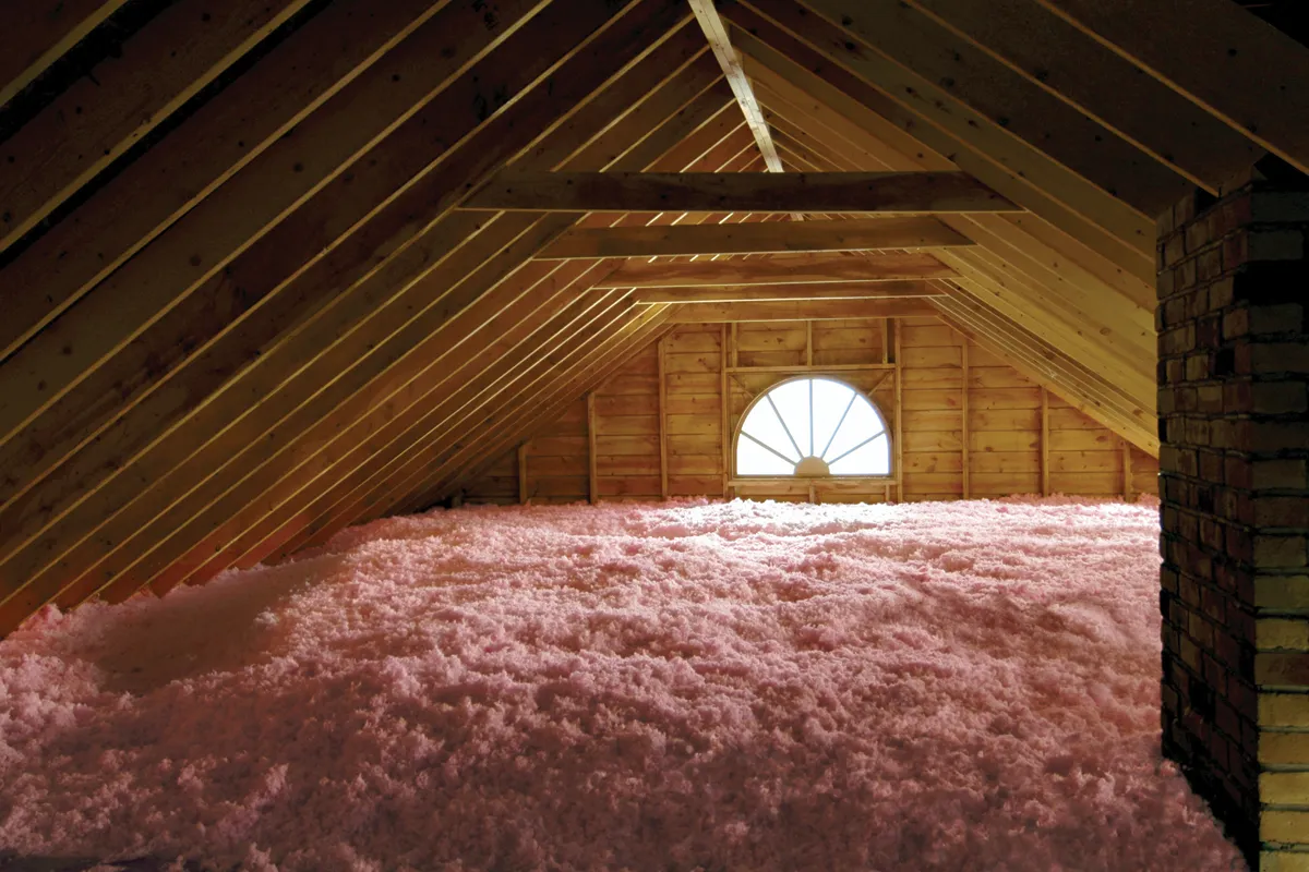 Attic space with wooden beams and a window, filled with pink fiberglass insulation material.