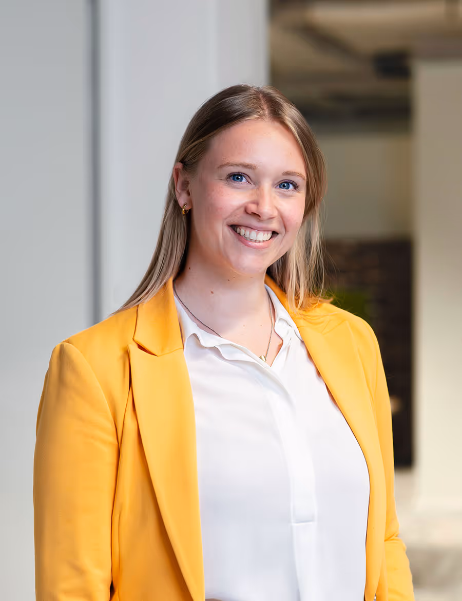 Jonge vrouw met lang haar en een lichtblauw overhemd glimlacht in een lichte, neutrale kamer.