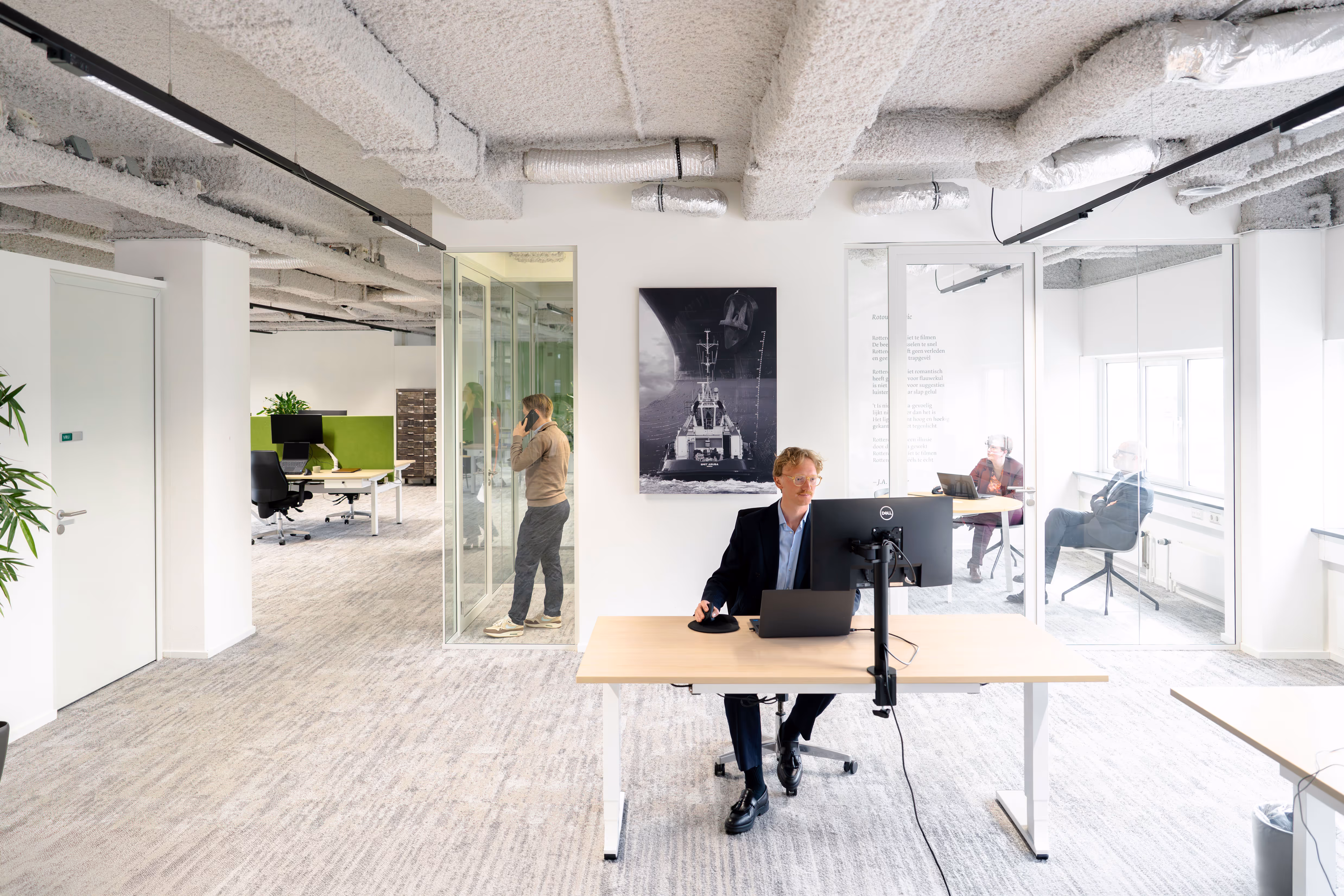Kantoorruimte met man aan bureau en anderen die in aparte ruimtes werken en praten.