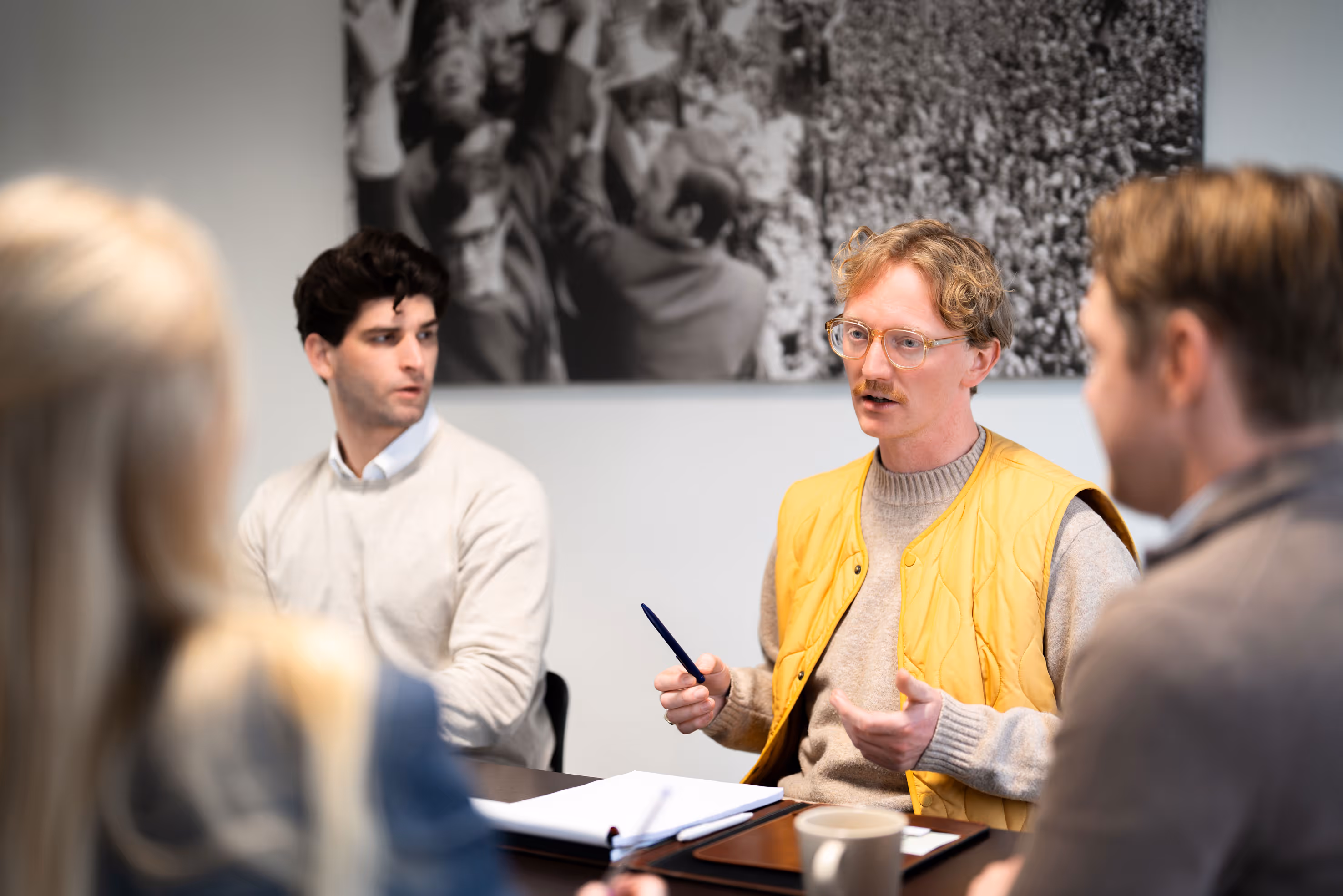 Man in yellow vest speaking to three colleagues at a meeting table with notebooks and a coffee mug.