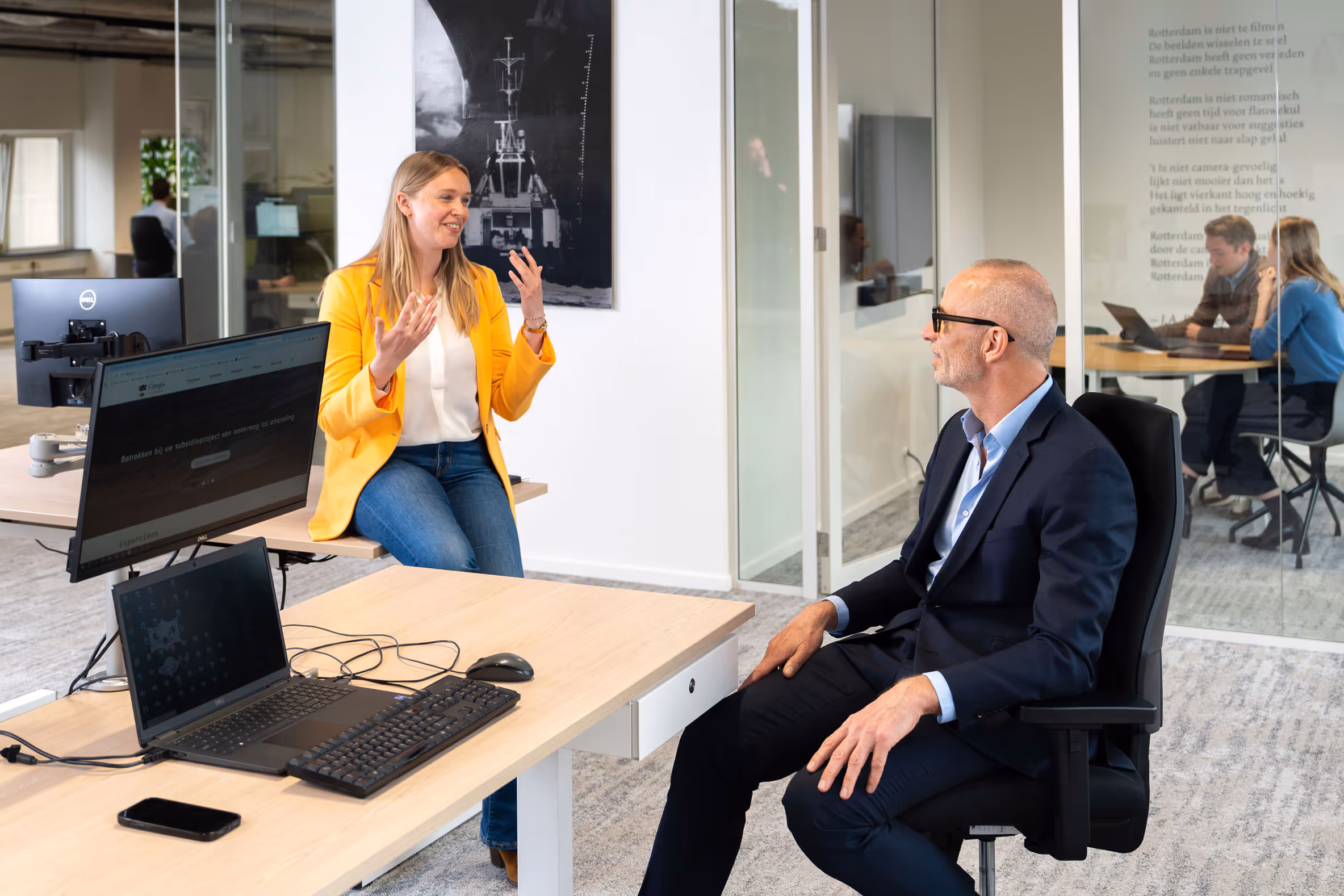 Vrouw in gele jas praat met man in pak aan bureau in modern kantoor.