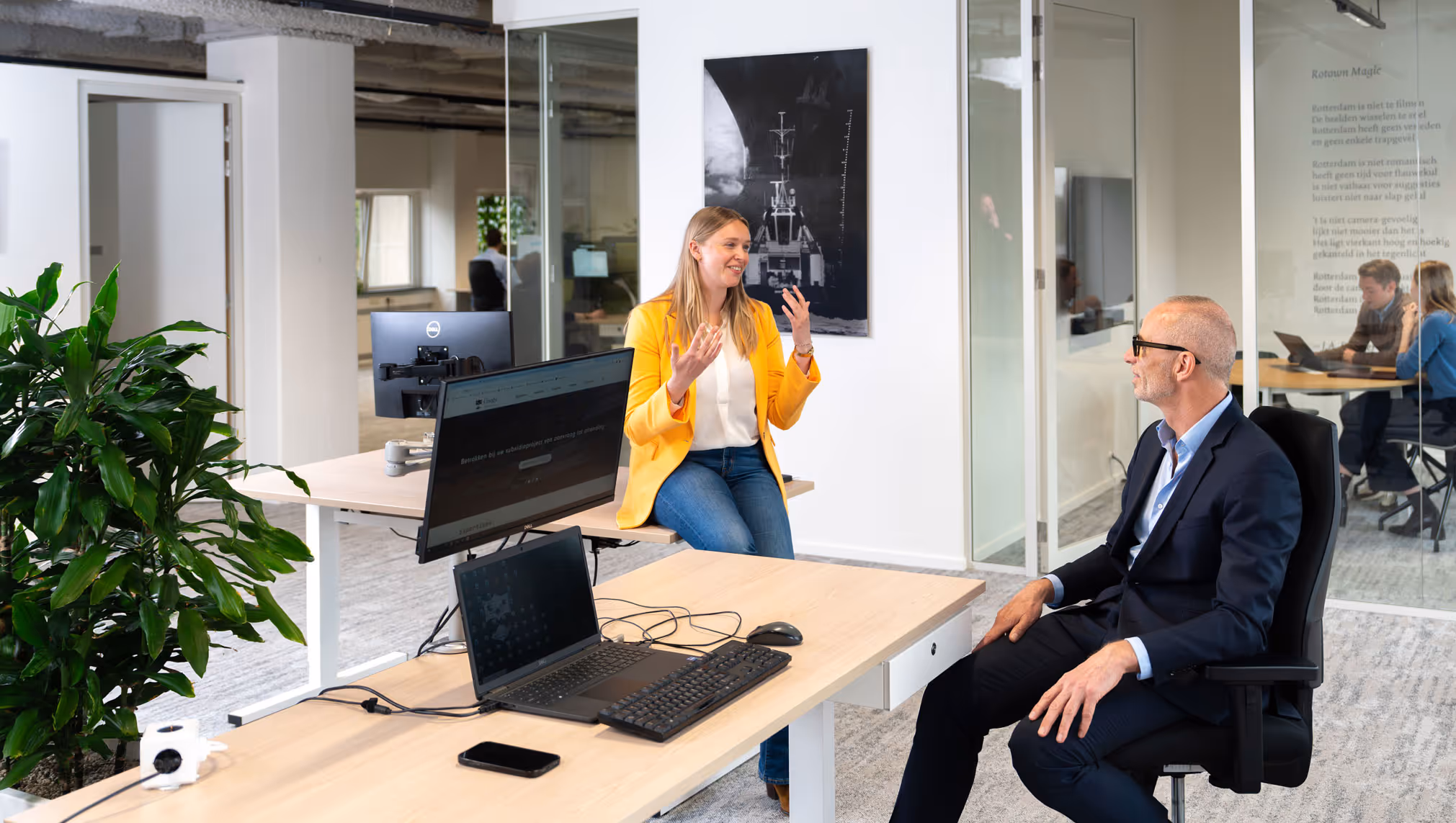 Twee collega’s hebben een gesprek in een modern kantoor met zit-sta-bureaus en glazen vergaderruimte.