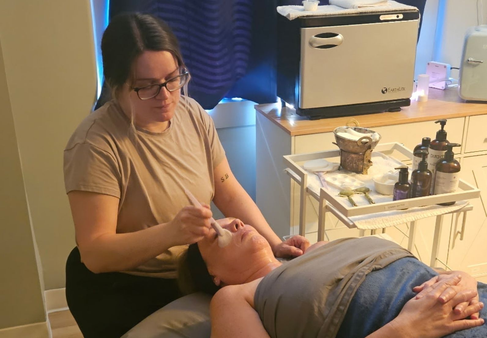 Woman wearing glasses applies facial treatment with a brush to a reclining client's face.