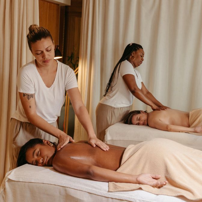 Two women receive back massages from massage therapists in a calm spa room with neutral tones.