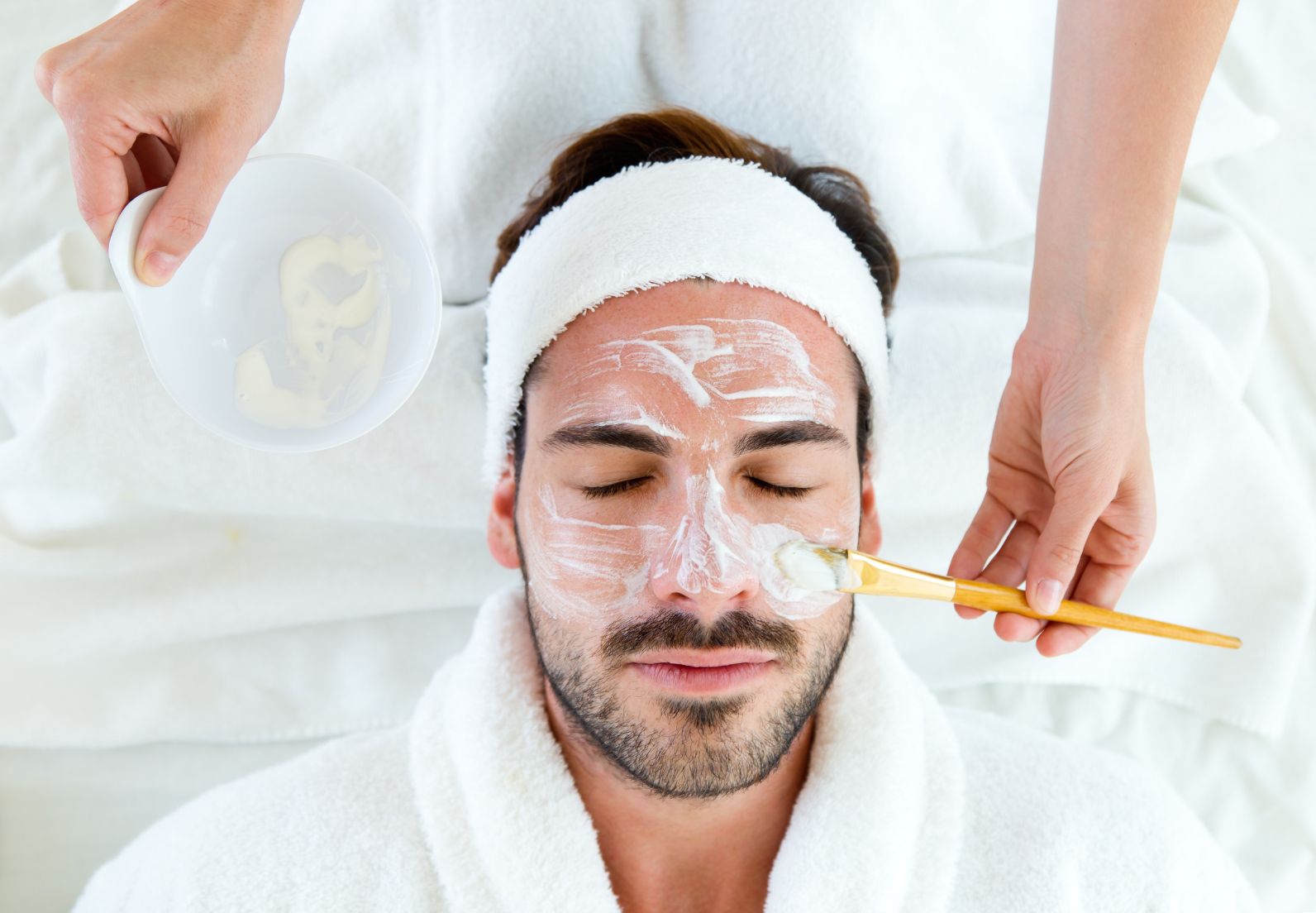 Man with eyes closed wearing a white headband having a facial mask applied with a brush at a spa.