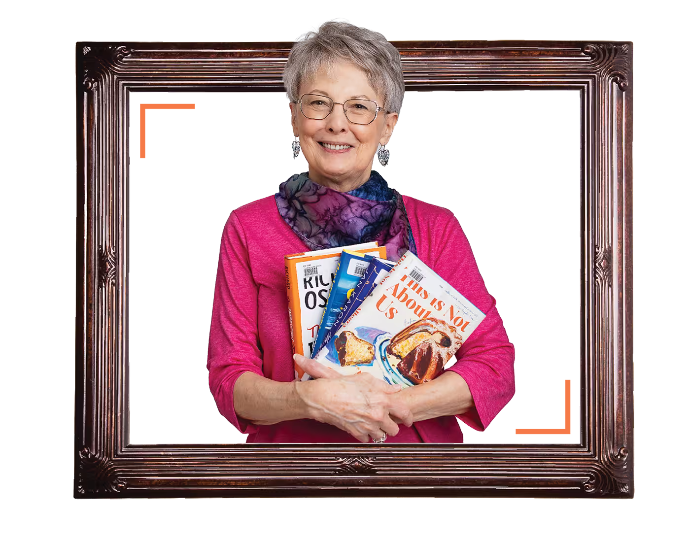 An older woman smiles while holding several cookbooks, posed within a large decorative frame.