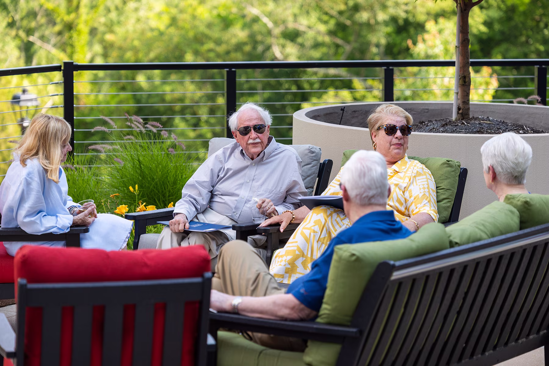 A group of seniors sitting outside on patio furniture, talking and enjoying a sunny day.
