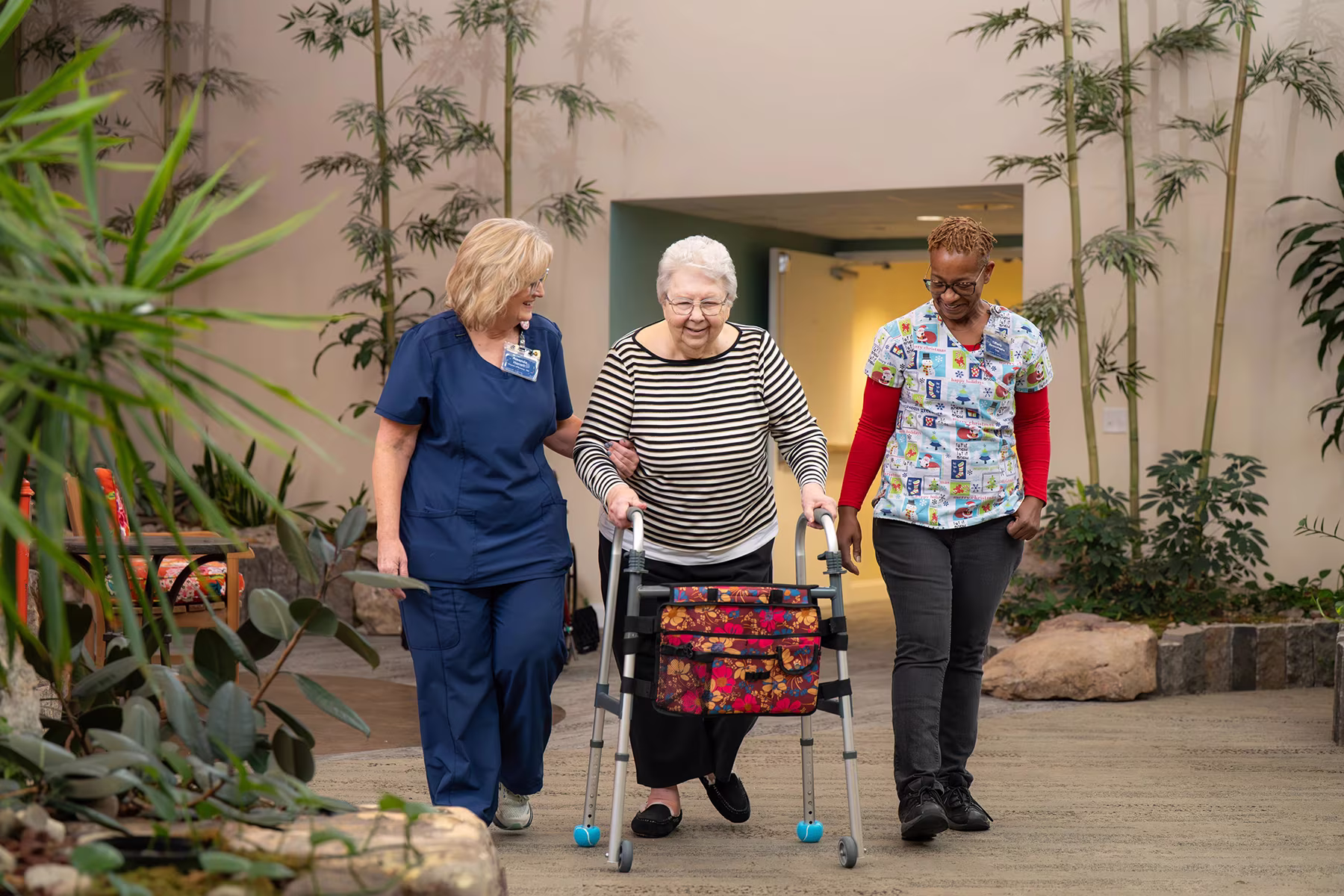 Two caregivers assist an elderly woman using a walker in a hallway with plants.