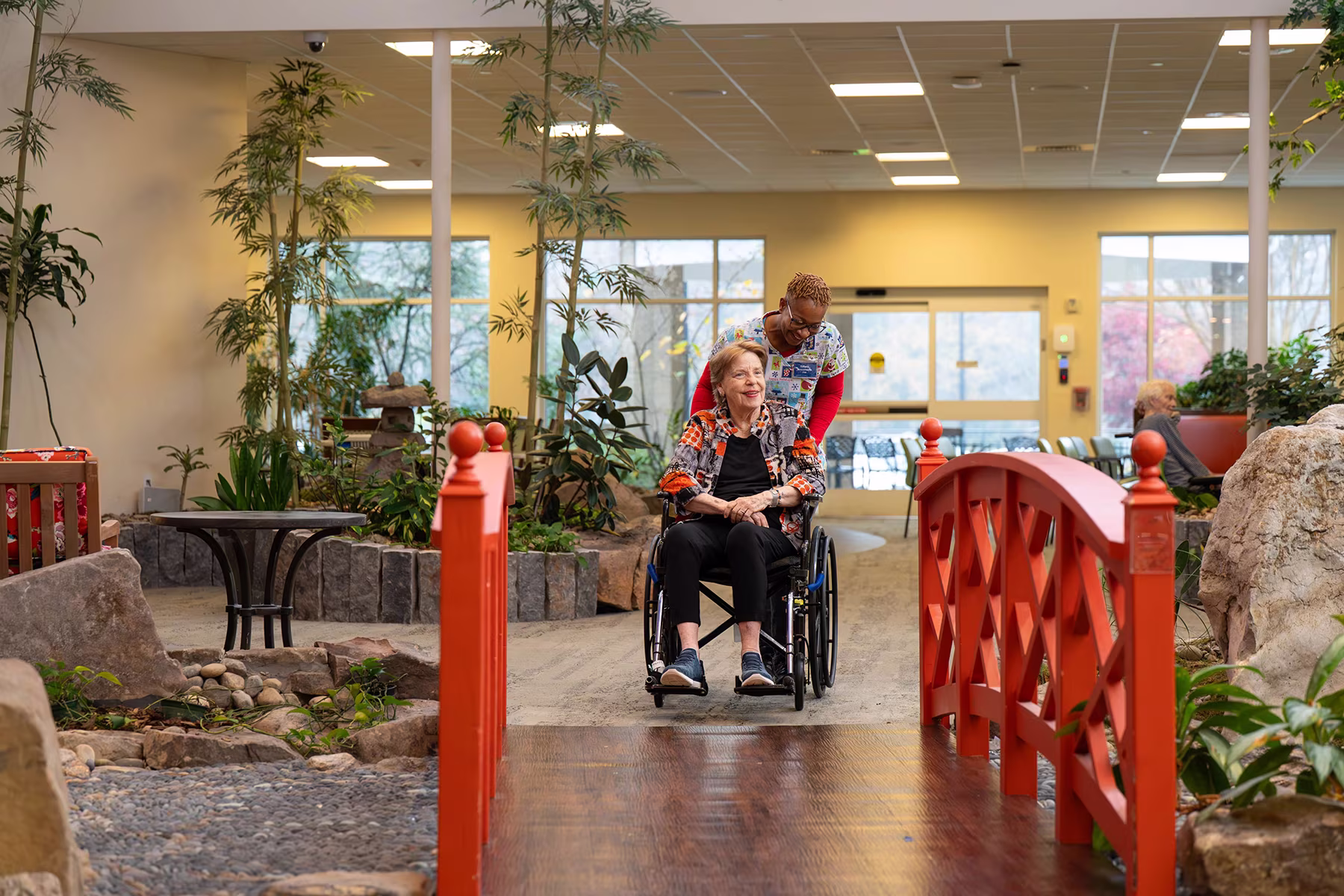 A woman pushes another woman in a wheelchair across a red bridge indoors.