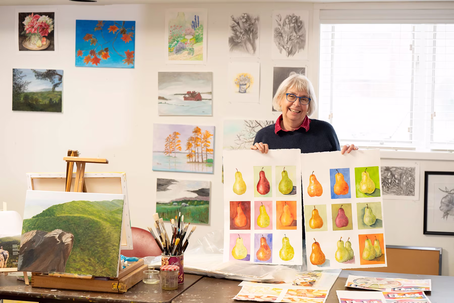 A person stands in an art studio, holding colorful artwork of pears.