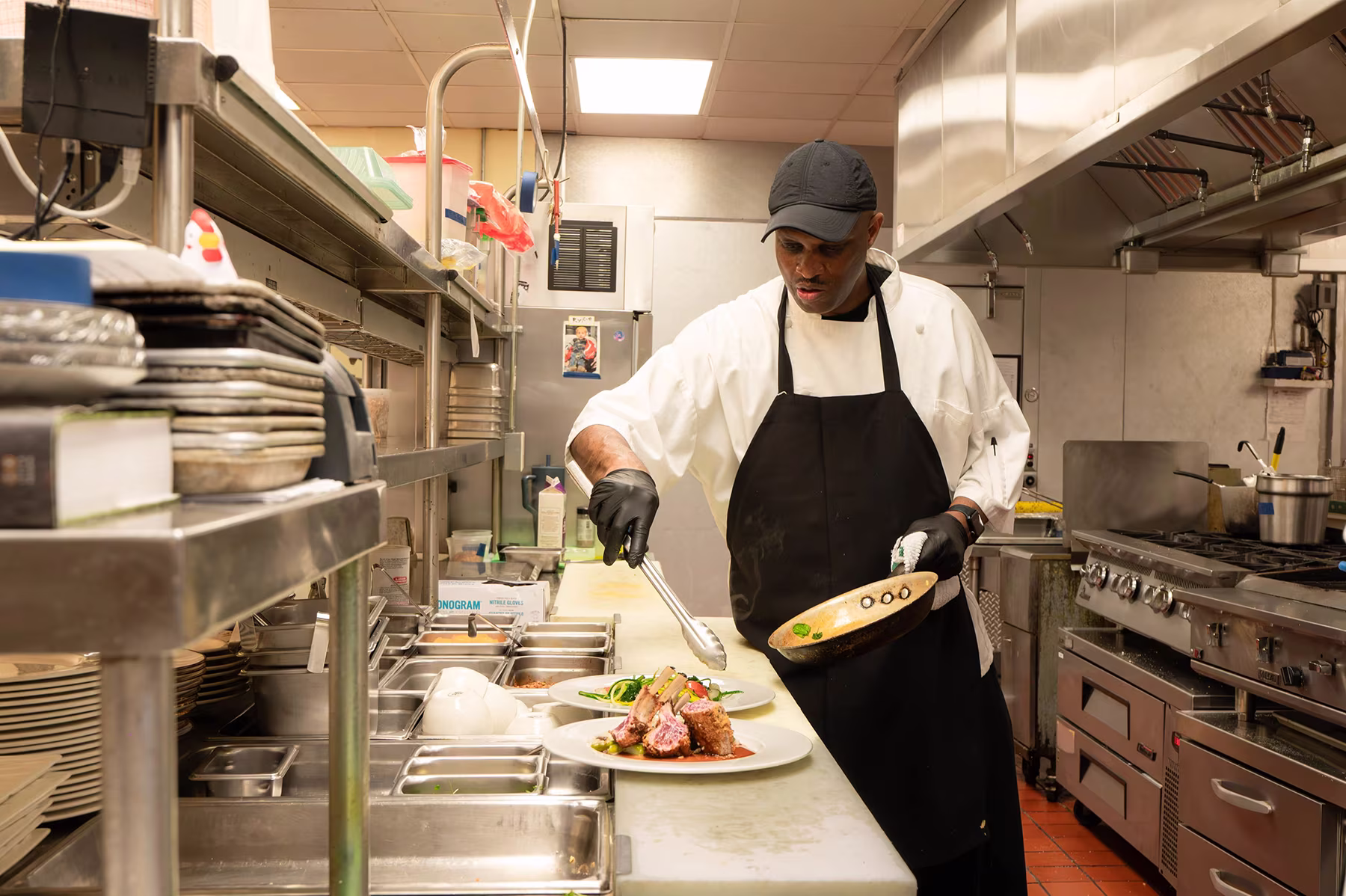 A chef in a kitchen carefully plating a dish with grilled meat and vegetables.