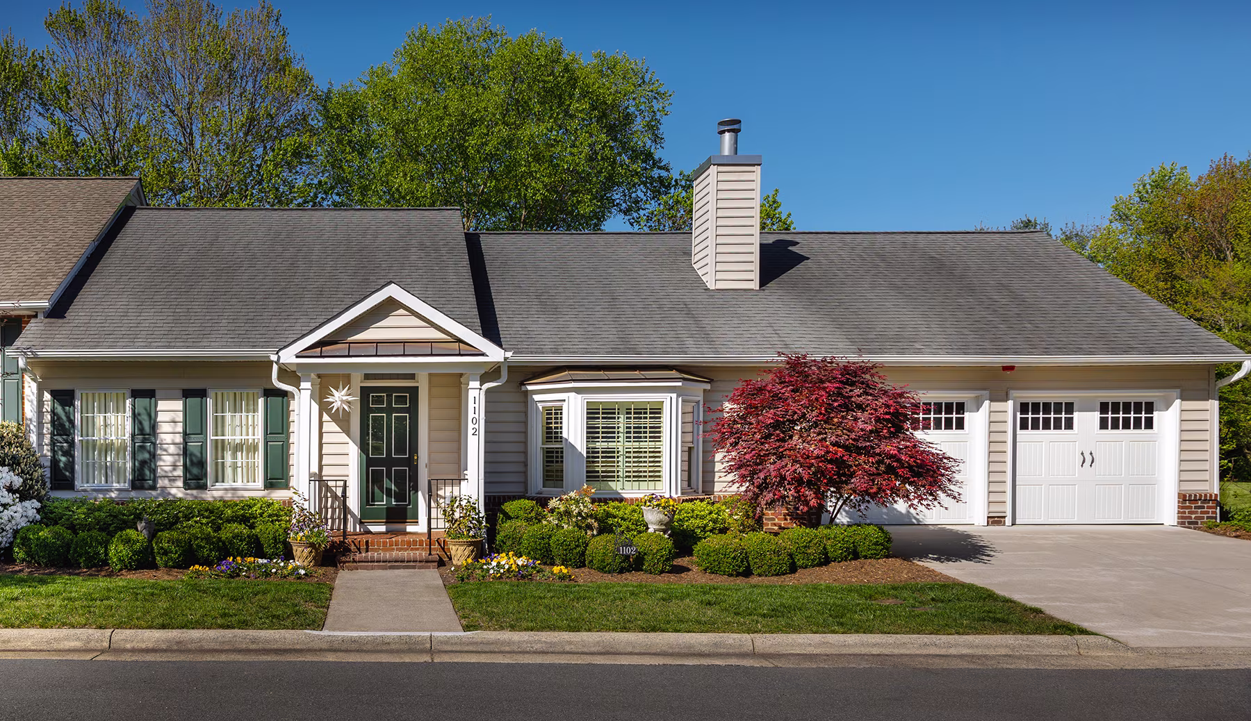 Single-story house with gray roof, white siding, and a small front garden with shrubs and a red tree.