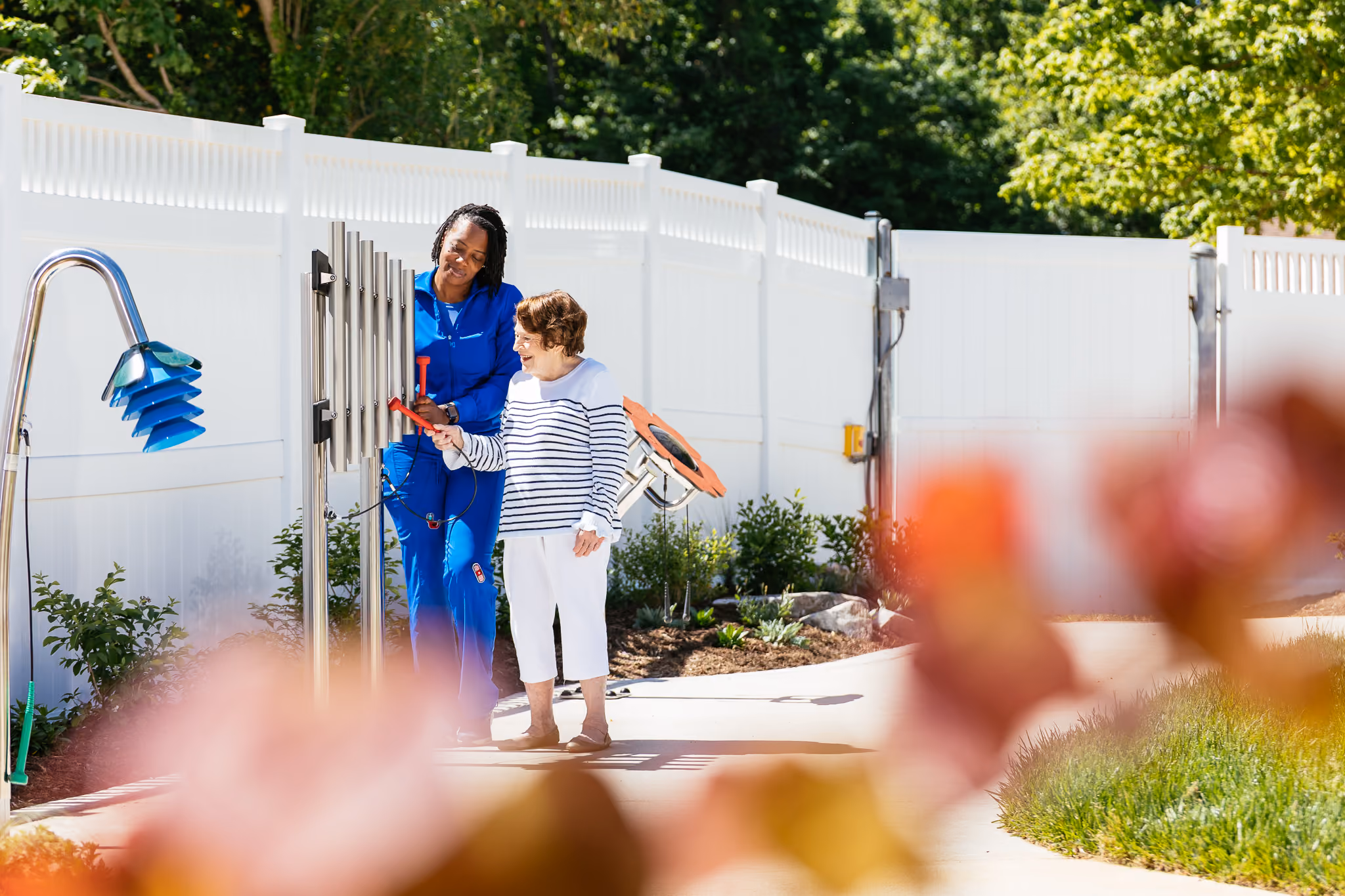 A caregiver assists a child in playing outdoor musical chimes along a garden walkway.