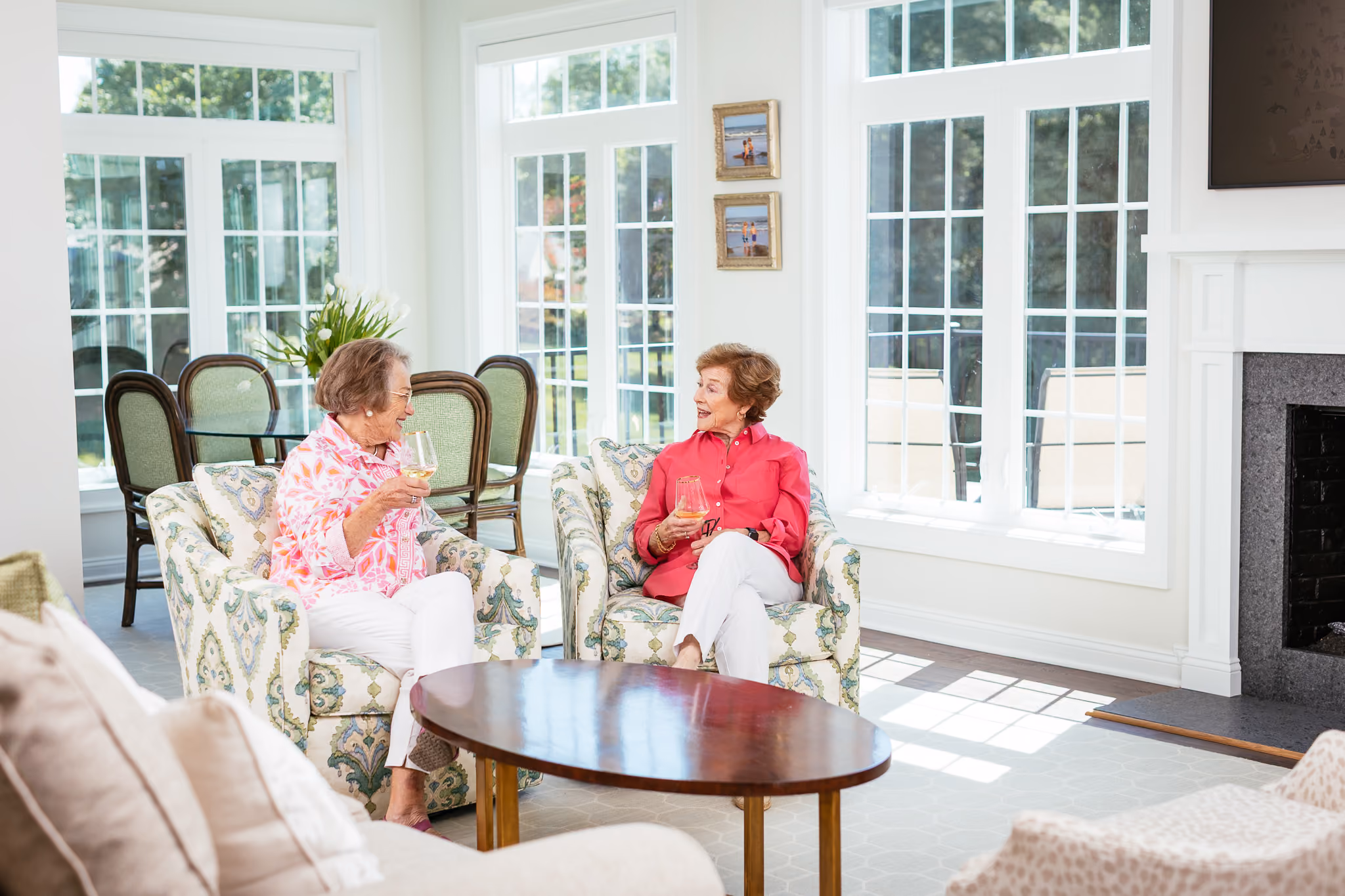 Two women sit on armchairs in a bright room, talking and holding glasses of wine.