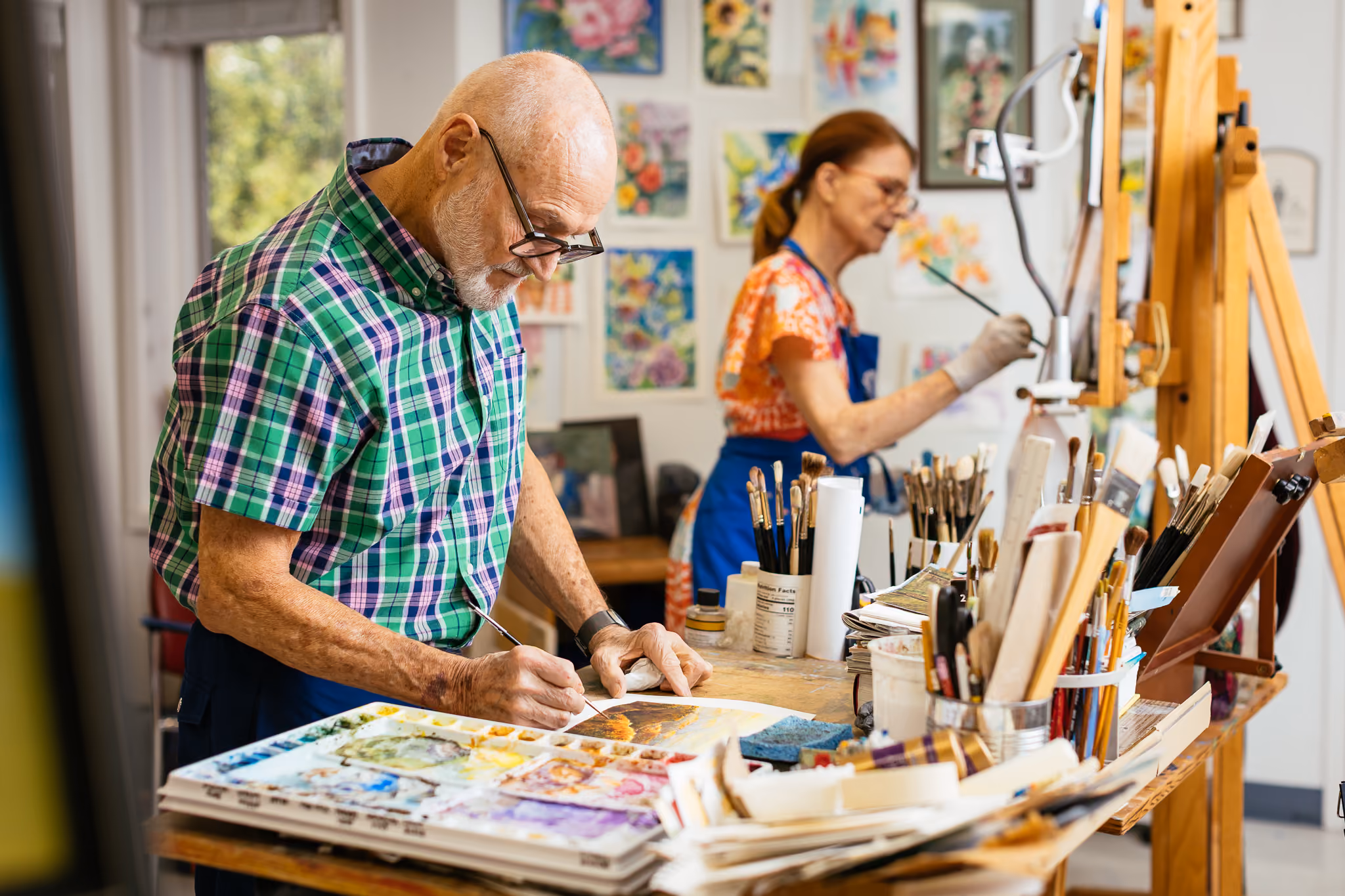 An elderly man paints at a table, while a woman works on a canvas in the background.