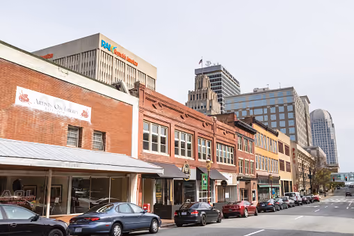 Street view of a city with parked cars and brick buildings under a cloudy sky.