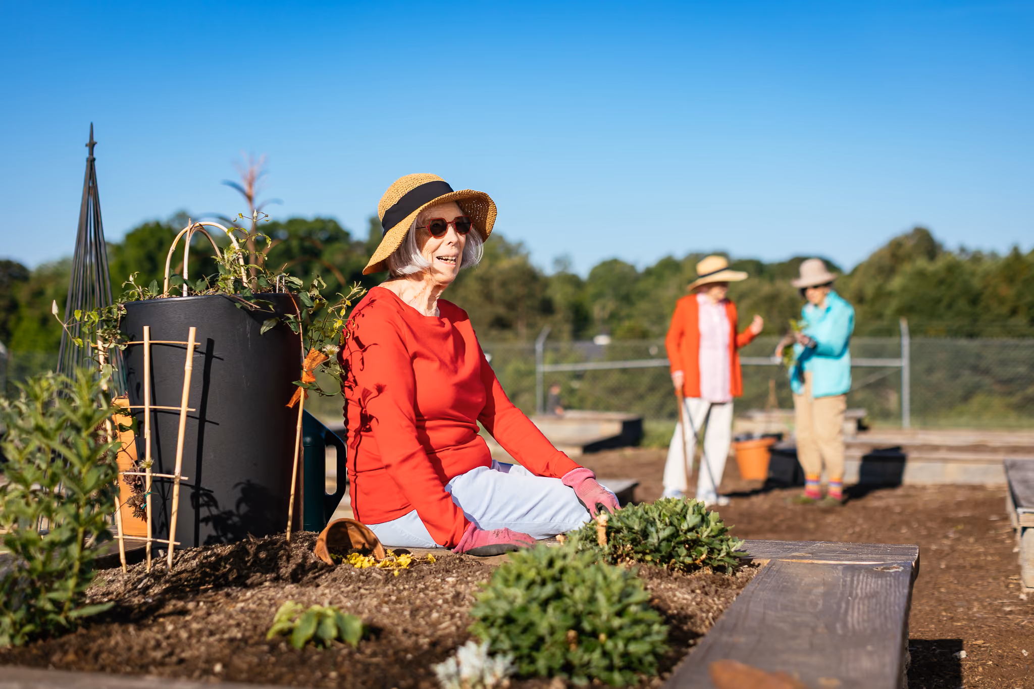 Elderly woman in a straw hat gardening, sitting by plants with two people chatting in the background.