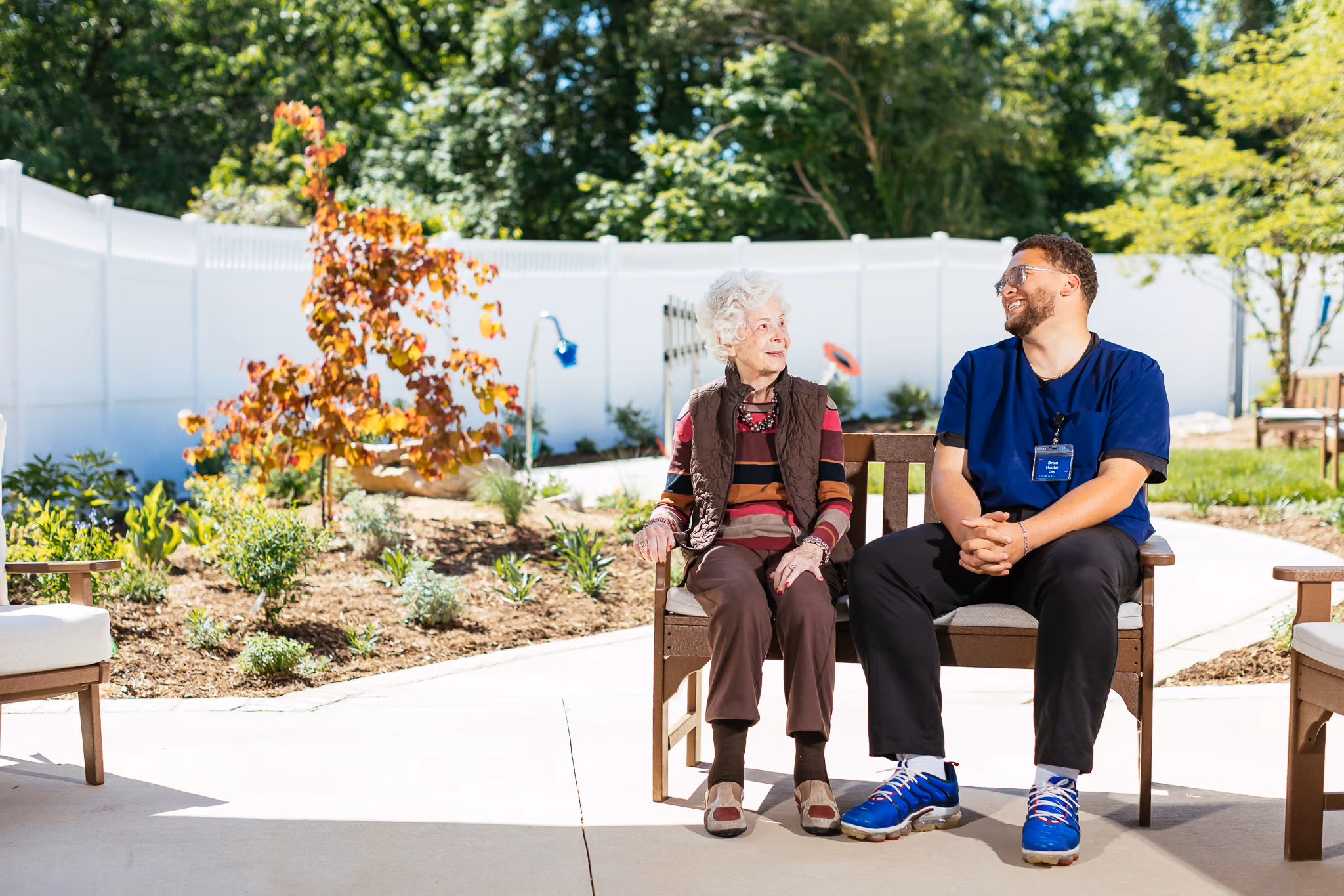 An elderly woman and a caregiver sit on a bench, smiling at each other in a garden.
