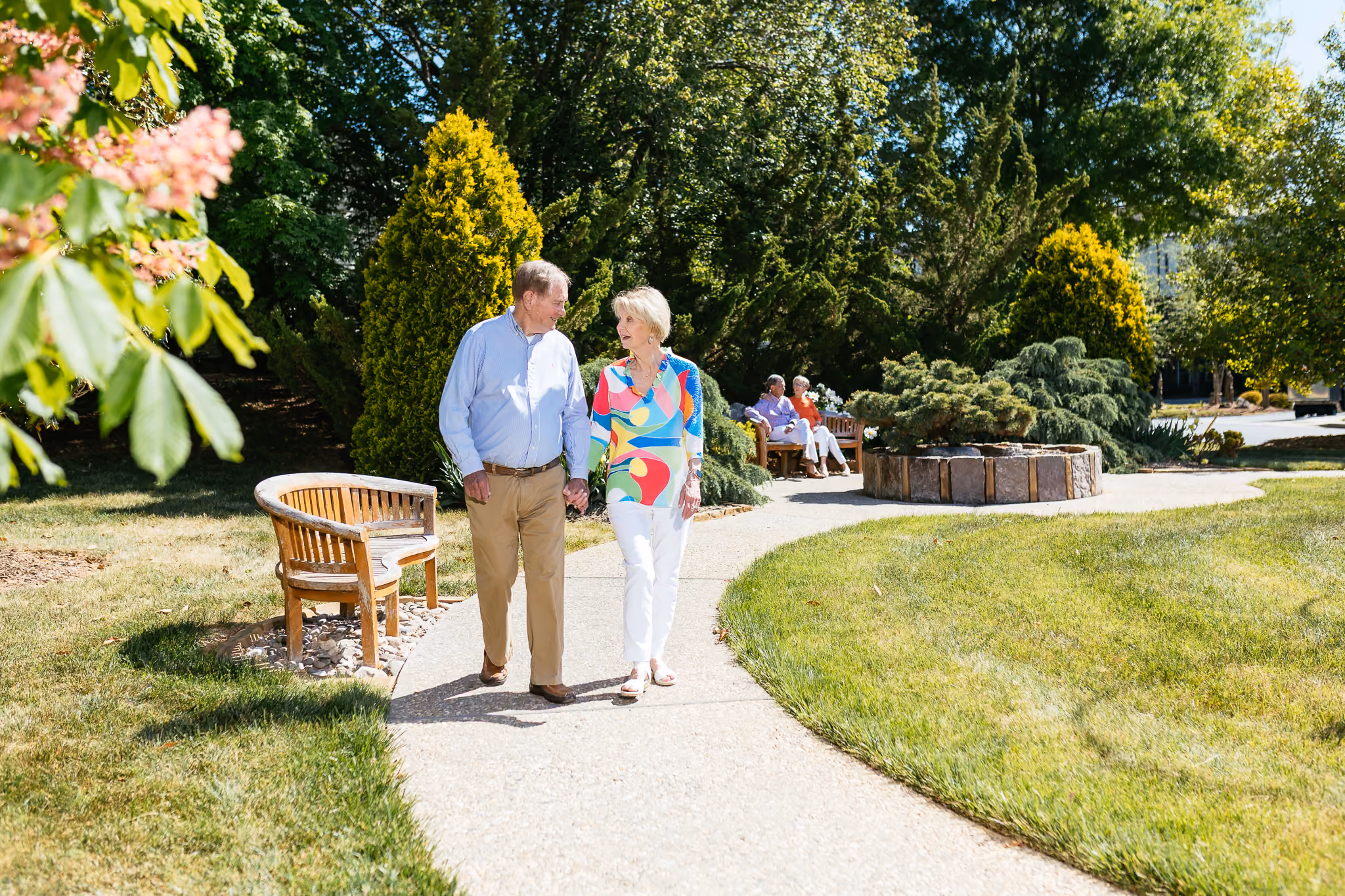 An elderly couple strolls together along a sunny path in a park, enjoying each other's company.