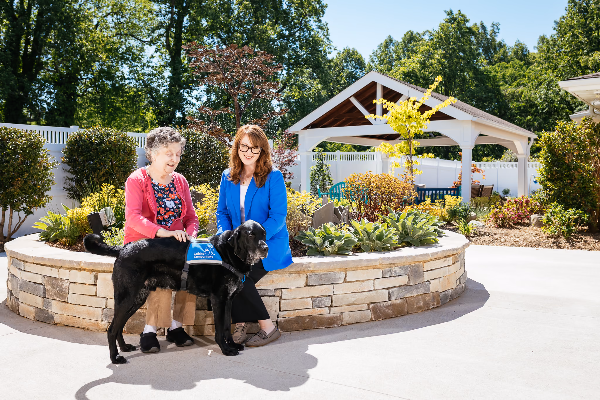 Two women sitting outside with a black service dog in front of a wooden gazebo.