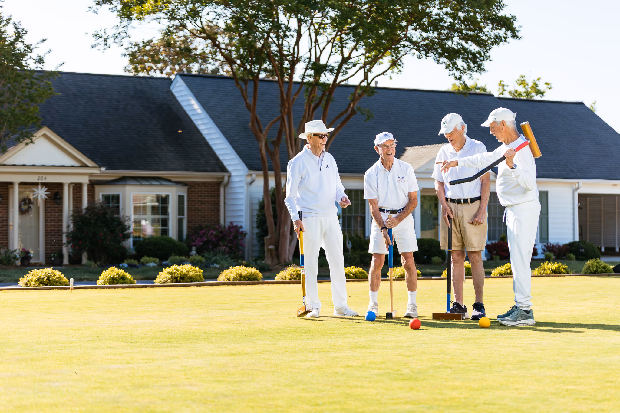 Four elderly men in white clothes play croquet on a lawn, smiling and talking.