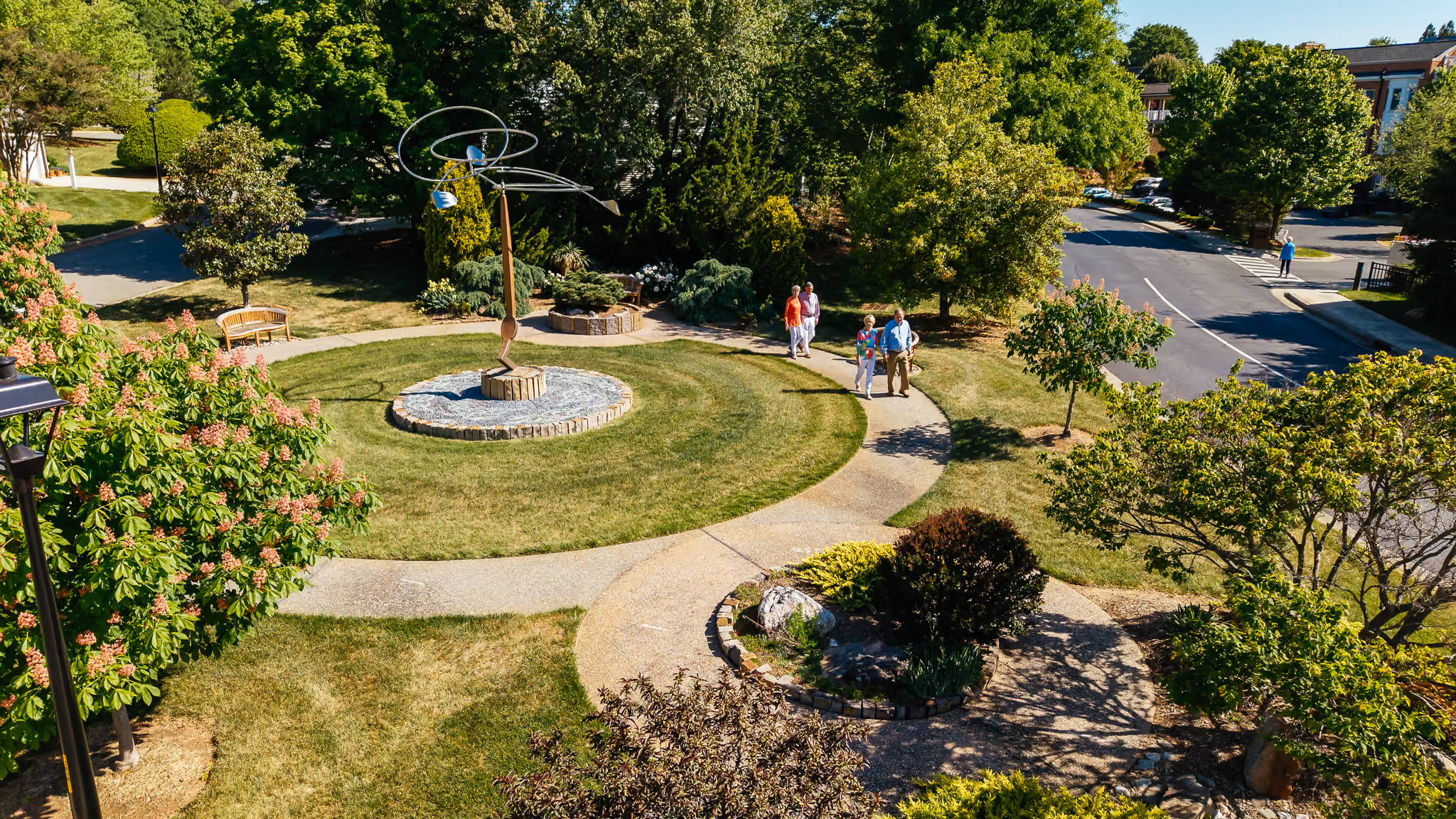 Two people walk along a path in a circular garden with a modern sculpture.