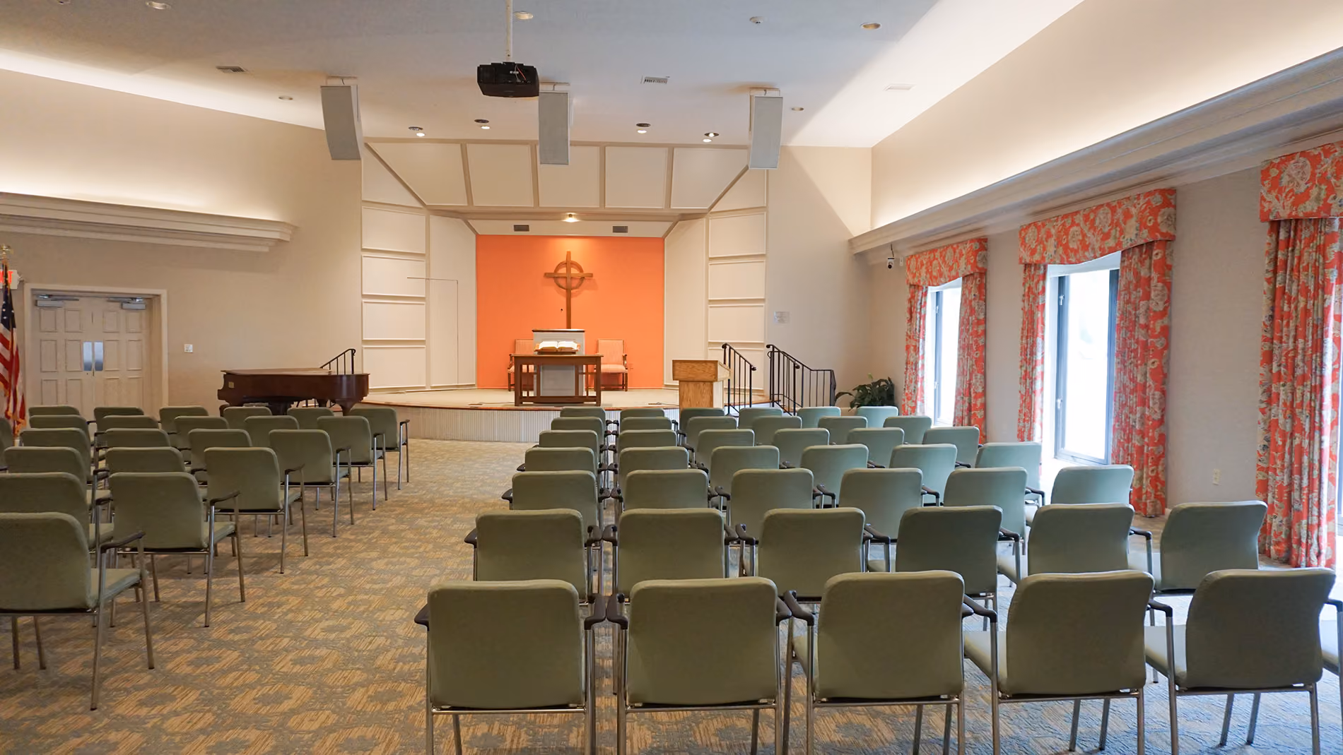 An empty church hall with rows of green chairs facing a stage with a lectern and cross.