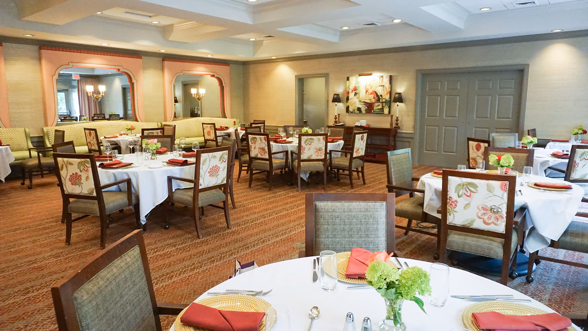 A dining room with neatly arranged tables, set with white tablecloths, red napkins, and floral chairs.