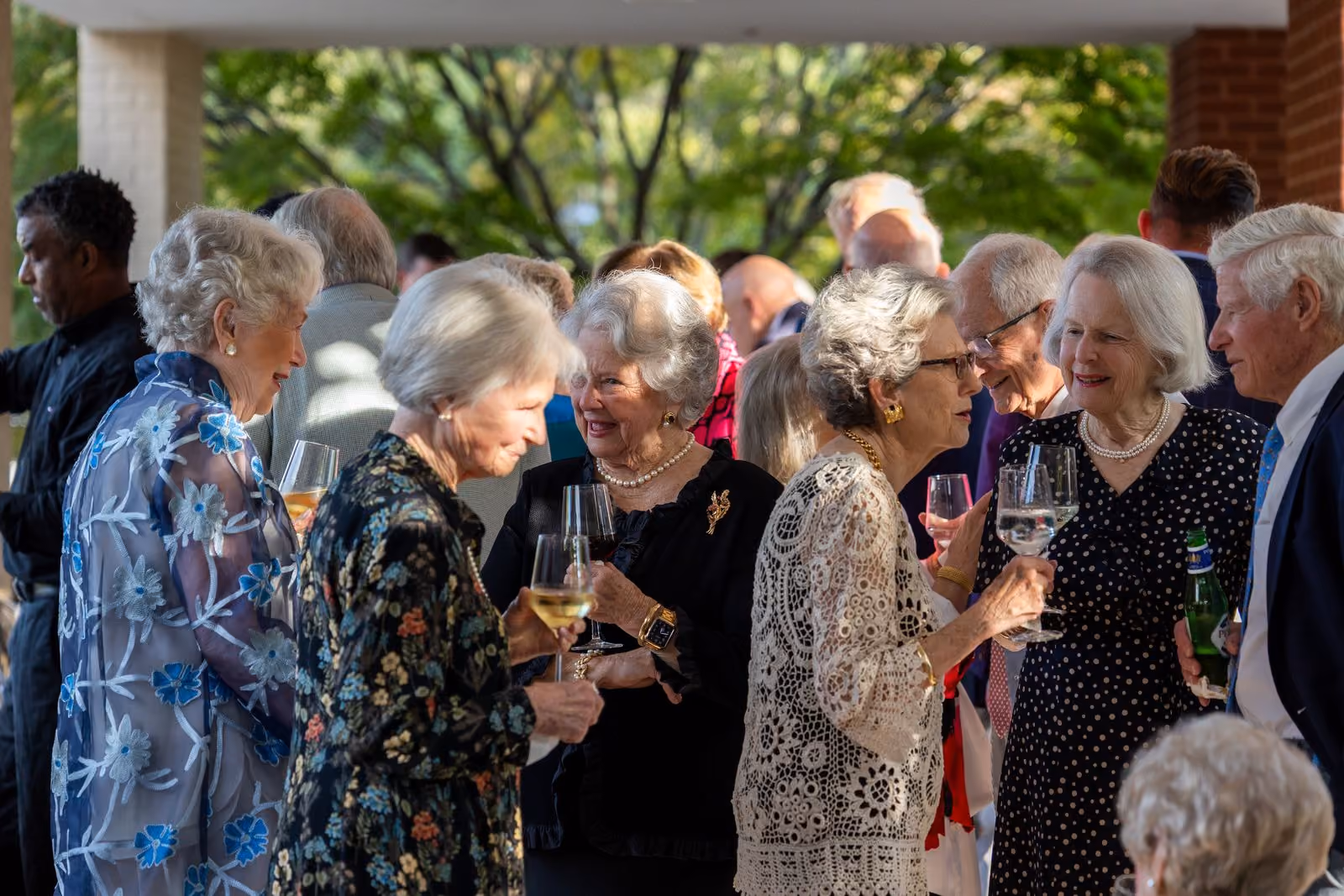 A group of elderly people are gathered, holding wine glasses and chatting at an outdoor event.