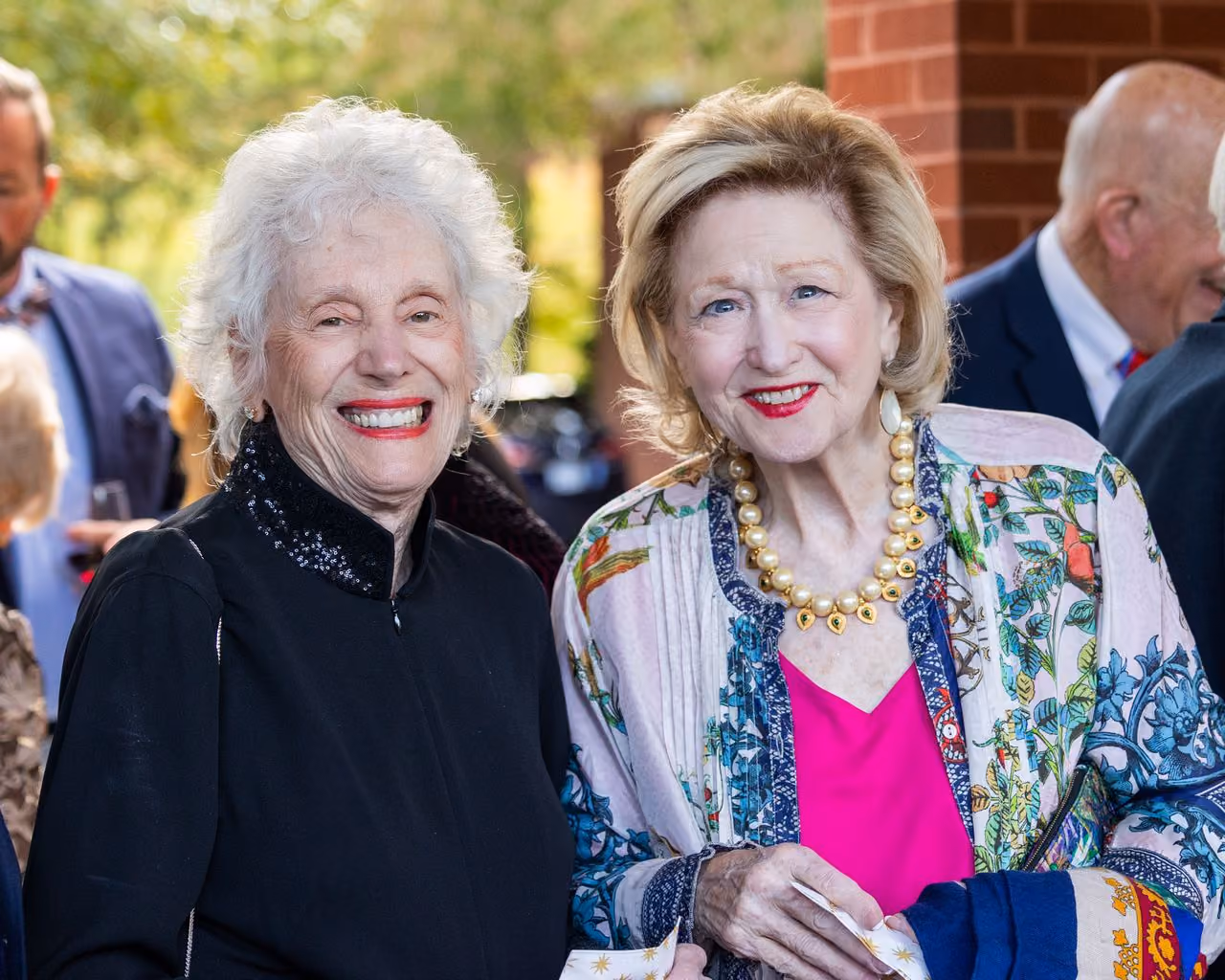 Two smiling women stand together, one in black and the other in a colorful outfit.