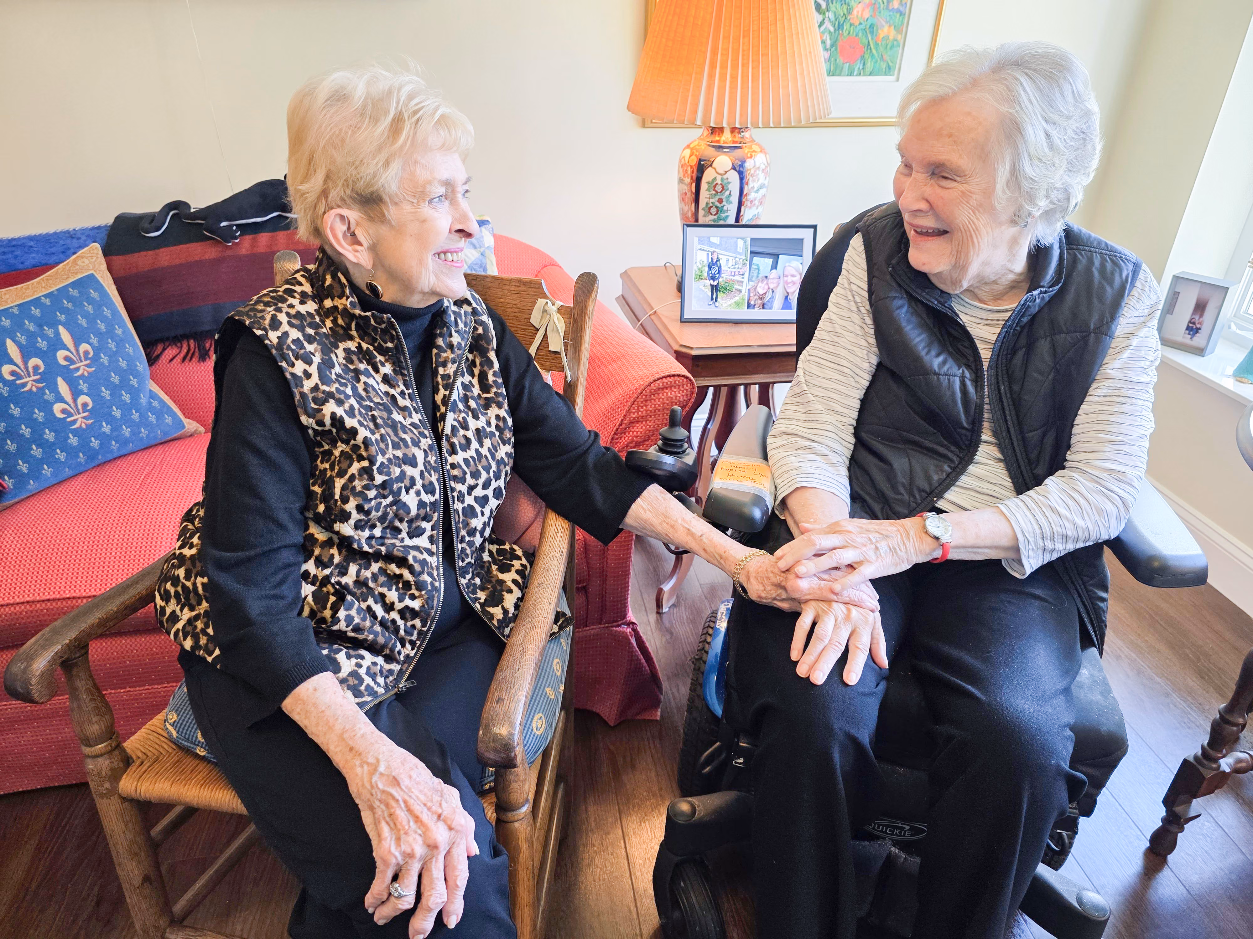 Two elderly women sit together, smiling and holding hands in a warmly lit living room.