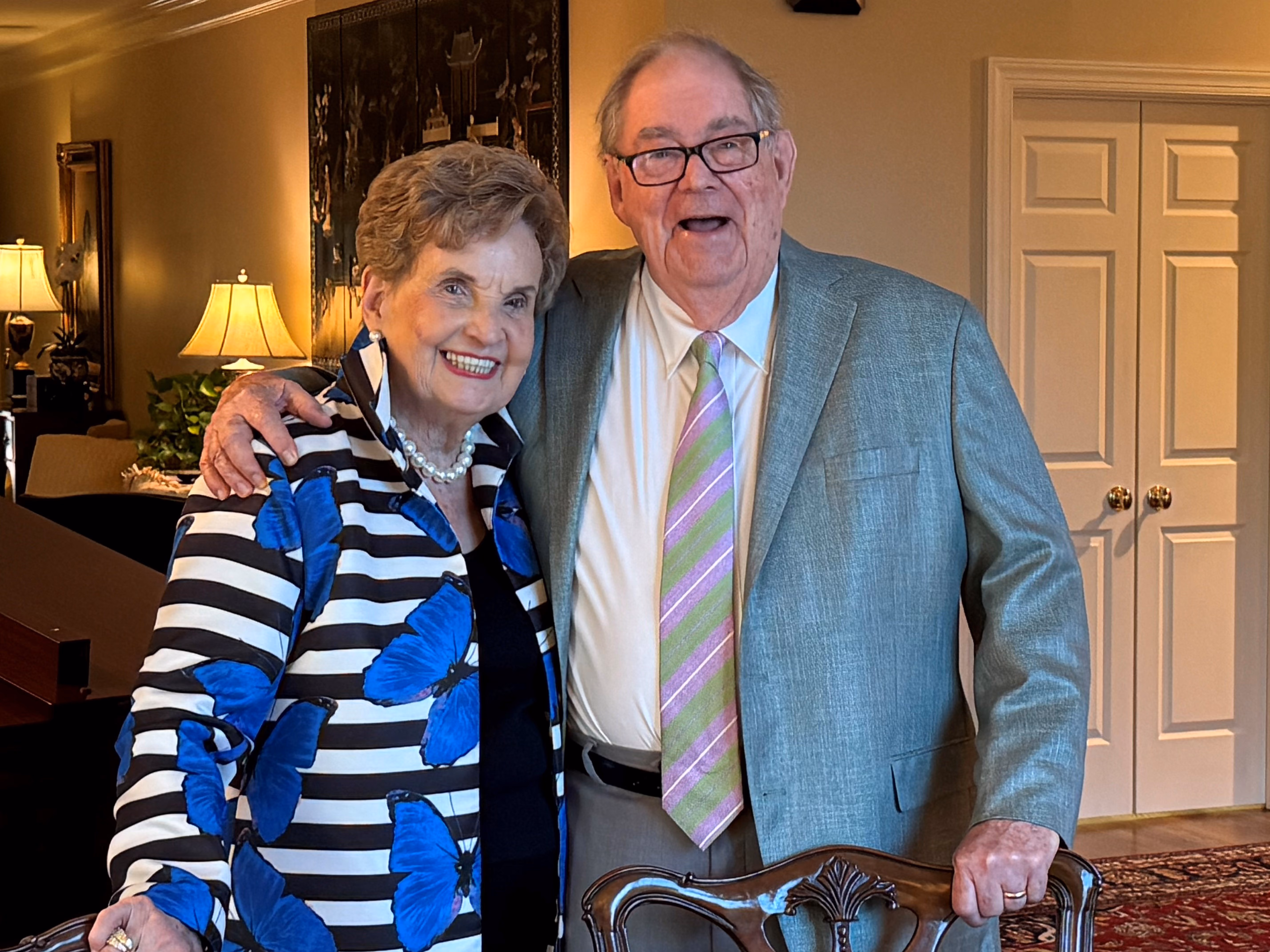 An older woman in a blue floral jacket stands smiling next to an older man in a suit.