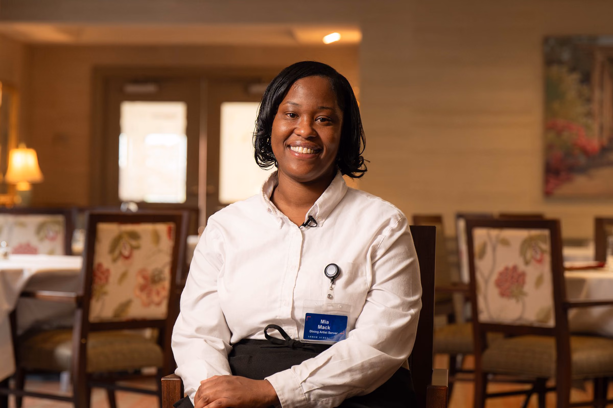 A person in a white shirt sits smiling in a dining room with chairs and tables.