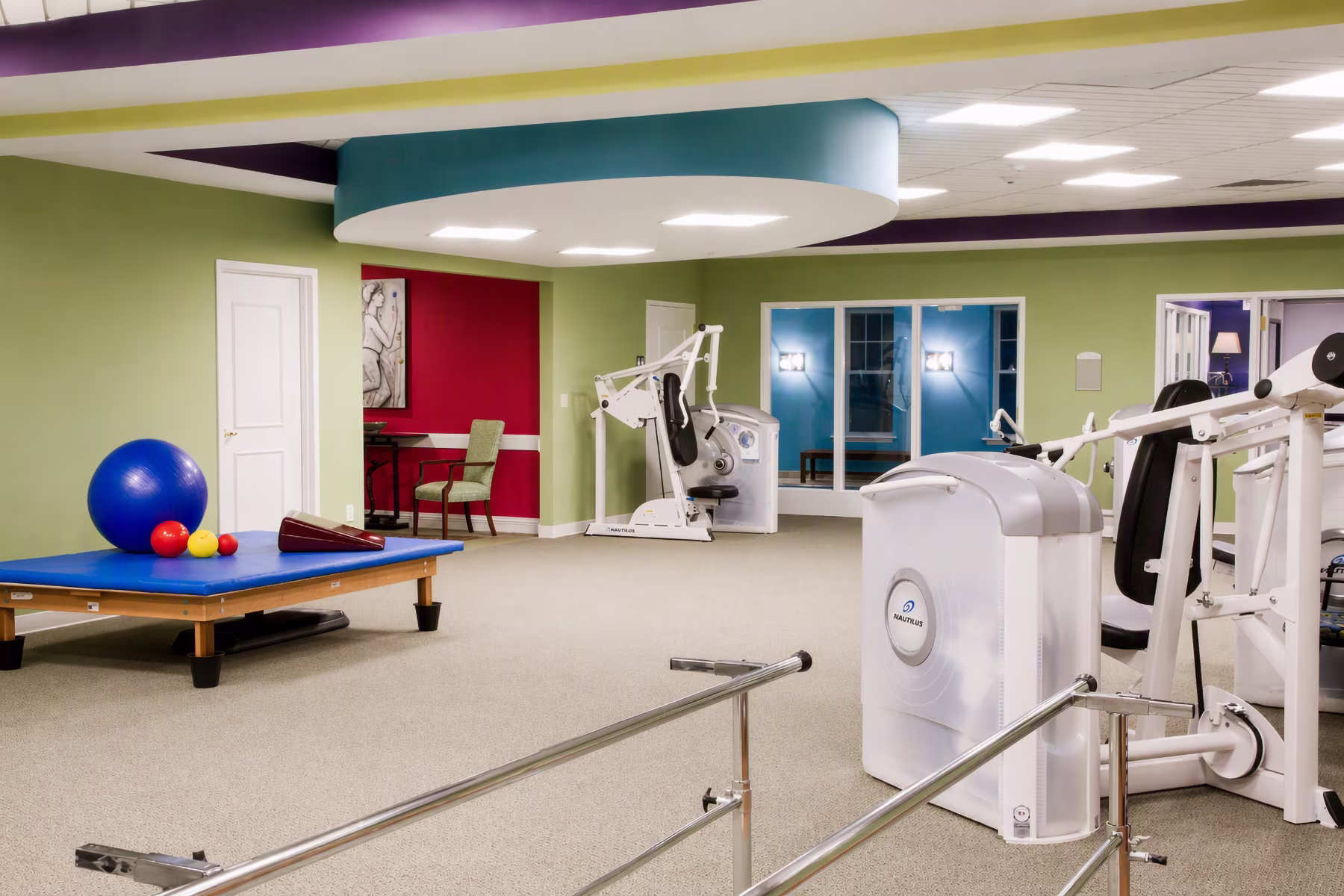 A fitness room with exercise machines, a padded table with therapy balls, and parallel bars.