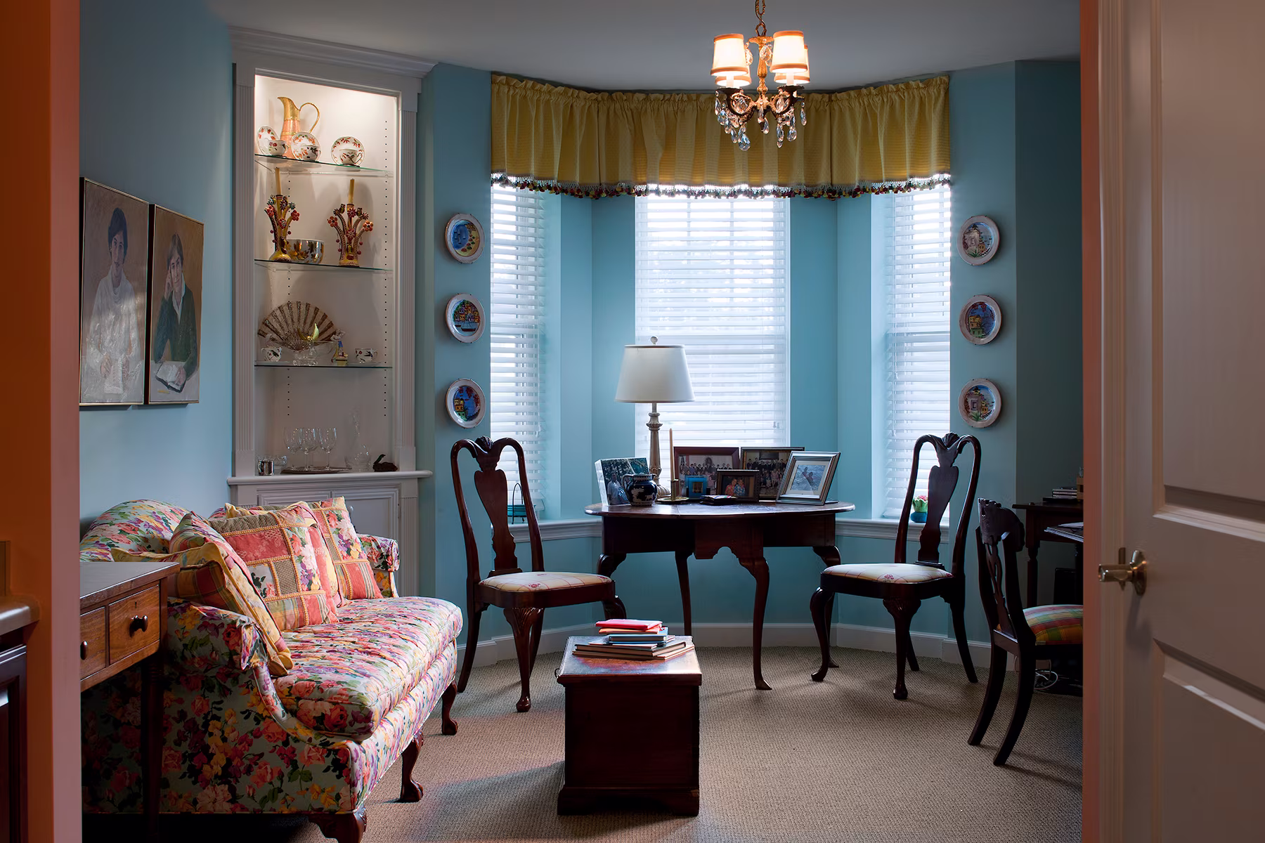 A cozy living room with a floral couch, wooden chairs, and a small table by the window.