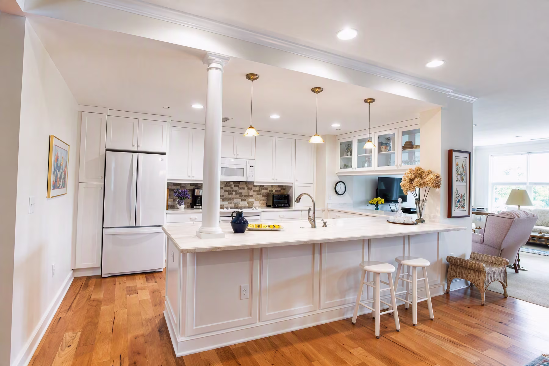 Spacious kitchen with white cabinets, marble island, and two white stools, brightened by pendant lights.
