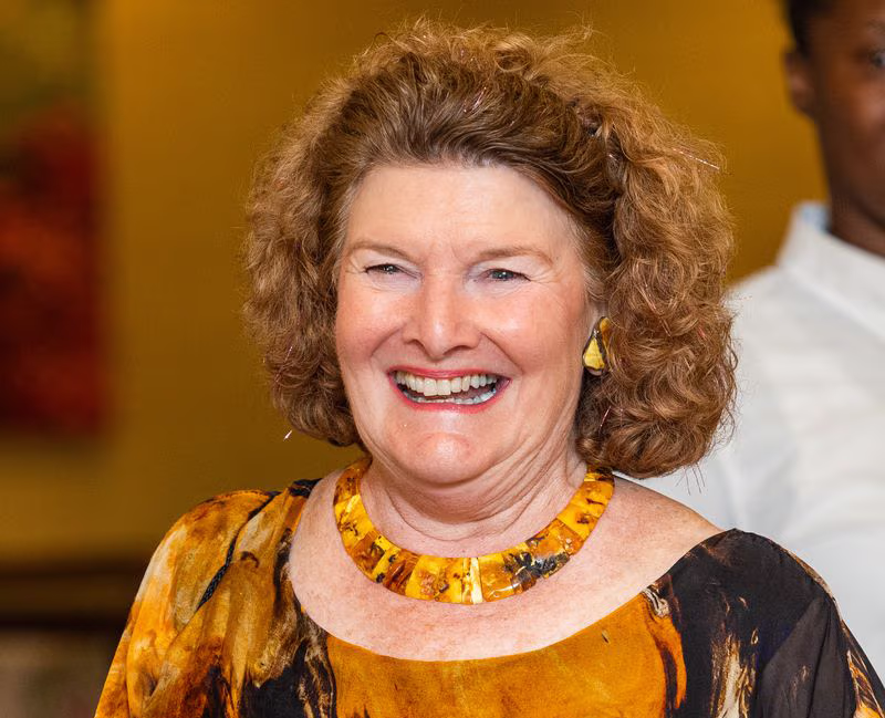 A woman with curly hair and a bright smile wears a colorful necklace and shirt.