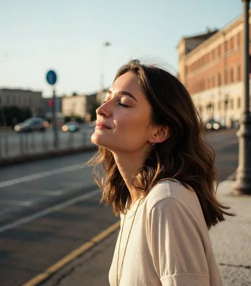 Woman with closed eyes and brown hair enjoying sunlight on a city street in the evening.