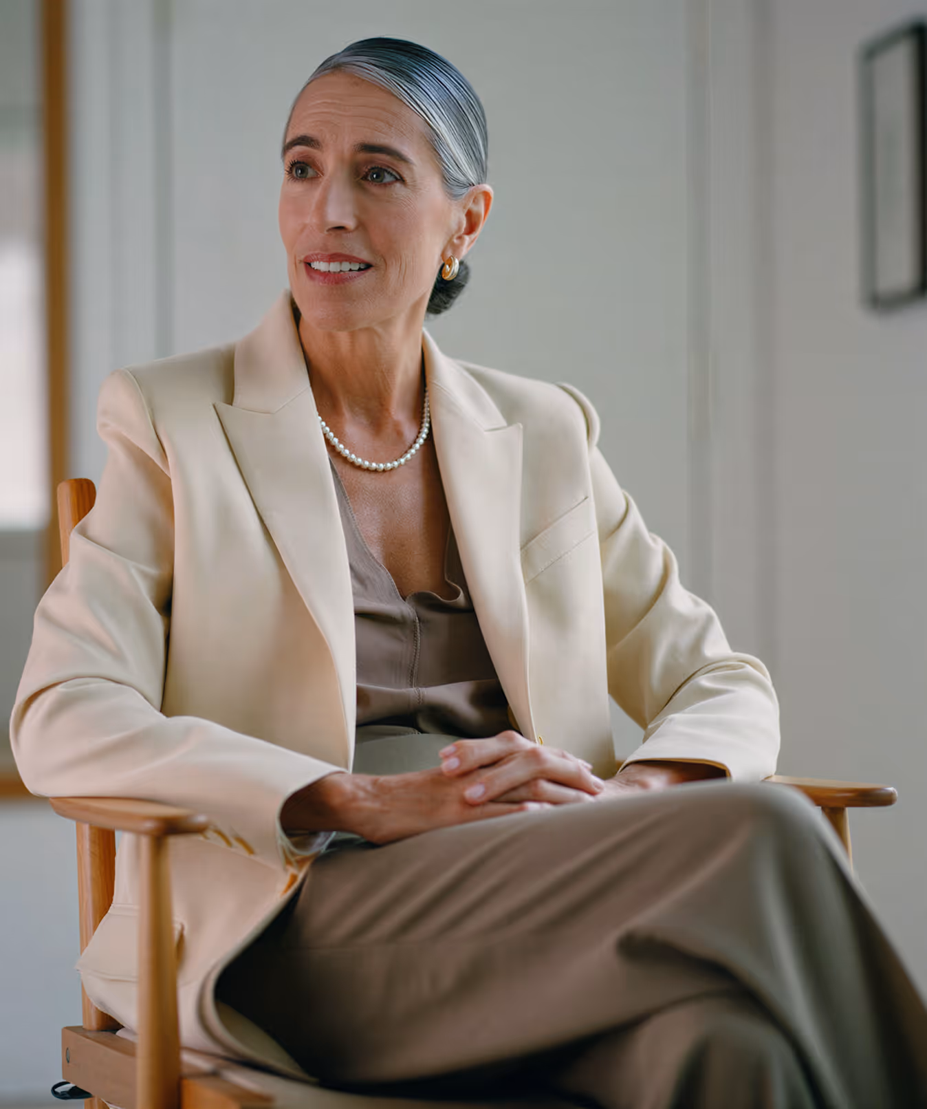 Elegant woman with silver hair in a cream blazer and pearl necklace sitting on a wooden chair.